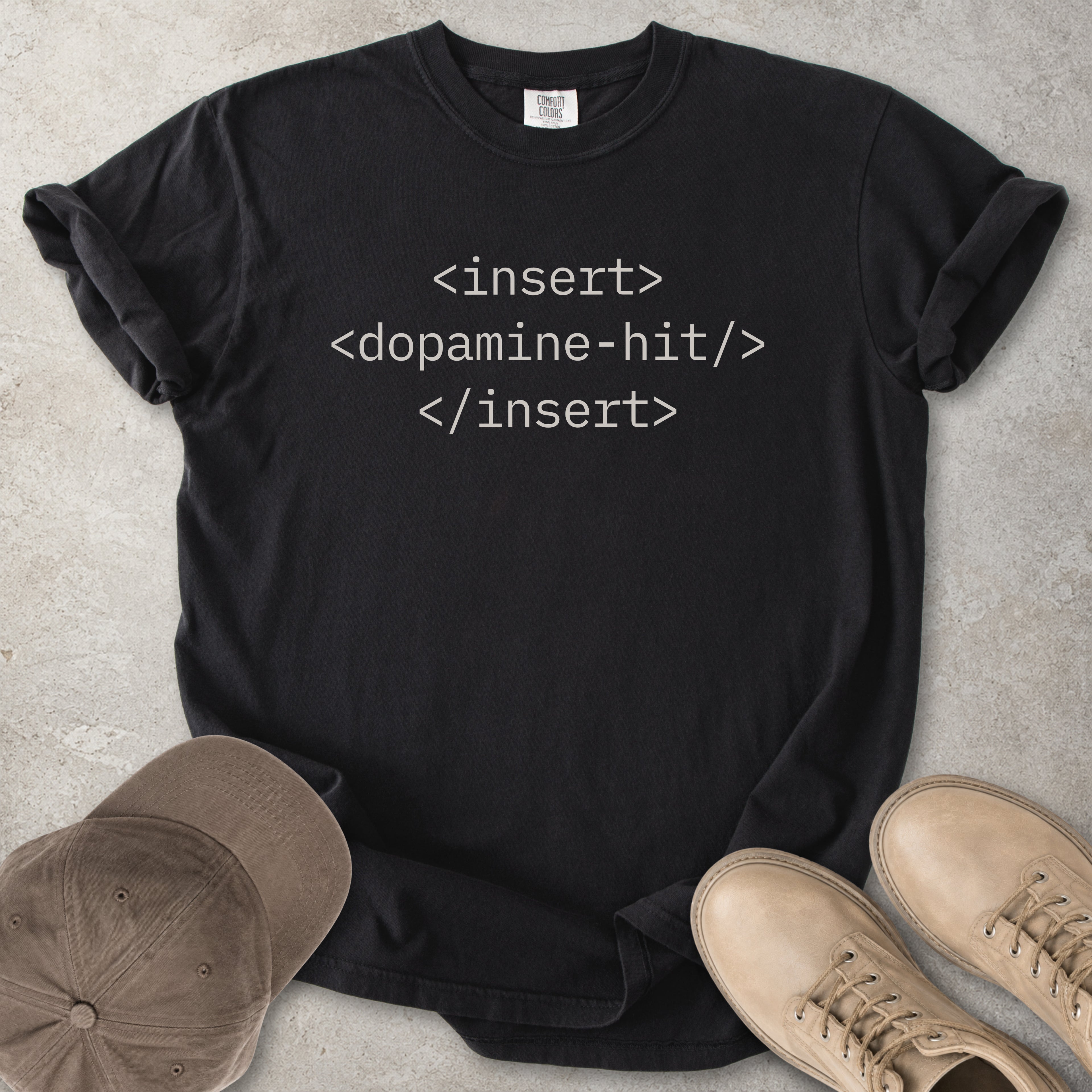 Black t-shirt with text 'insert <dopamine-hit> </insert>' on a light gray surface, surrounded by a cap and shoes.