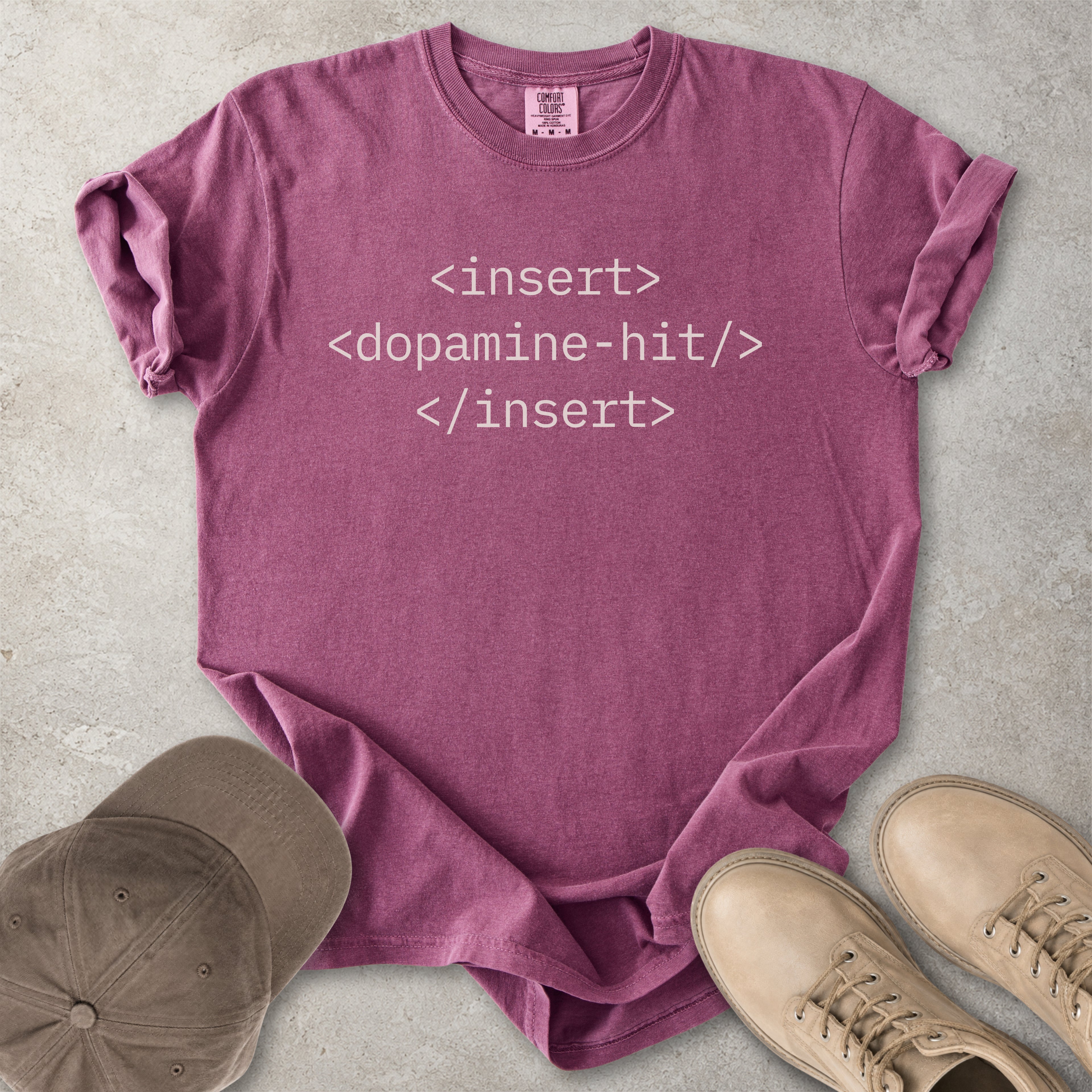 Purple t-shirt with text 'insert <dopamine-hit> </insert>' on a concrete surface with a cap and shoes.