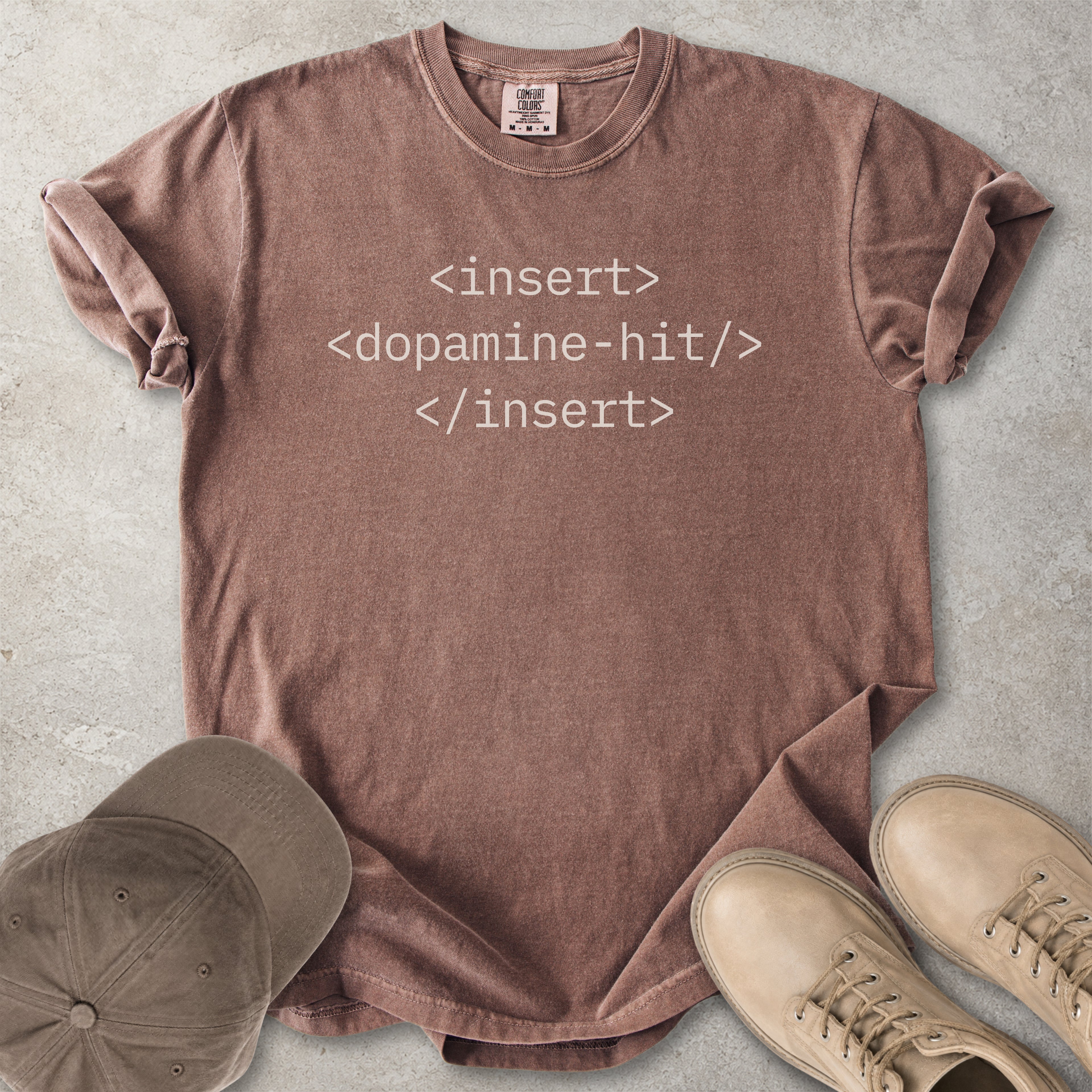 Brown t-shirt with text 'insert <dopamine-hit> </insert>' on a concrete surface, accompanied by a cap and shoes.