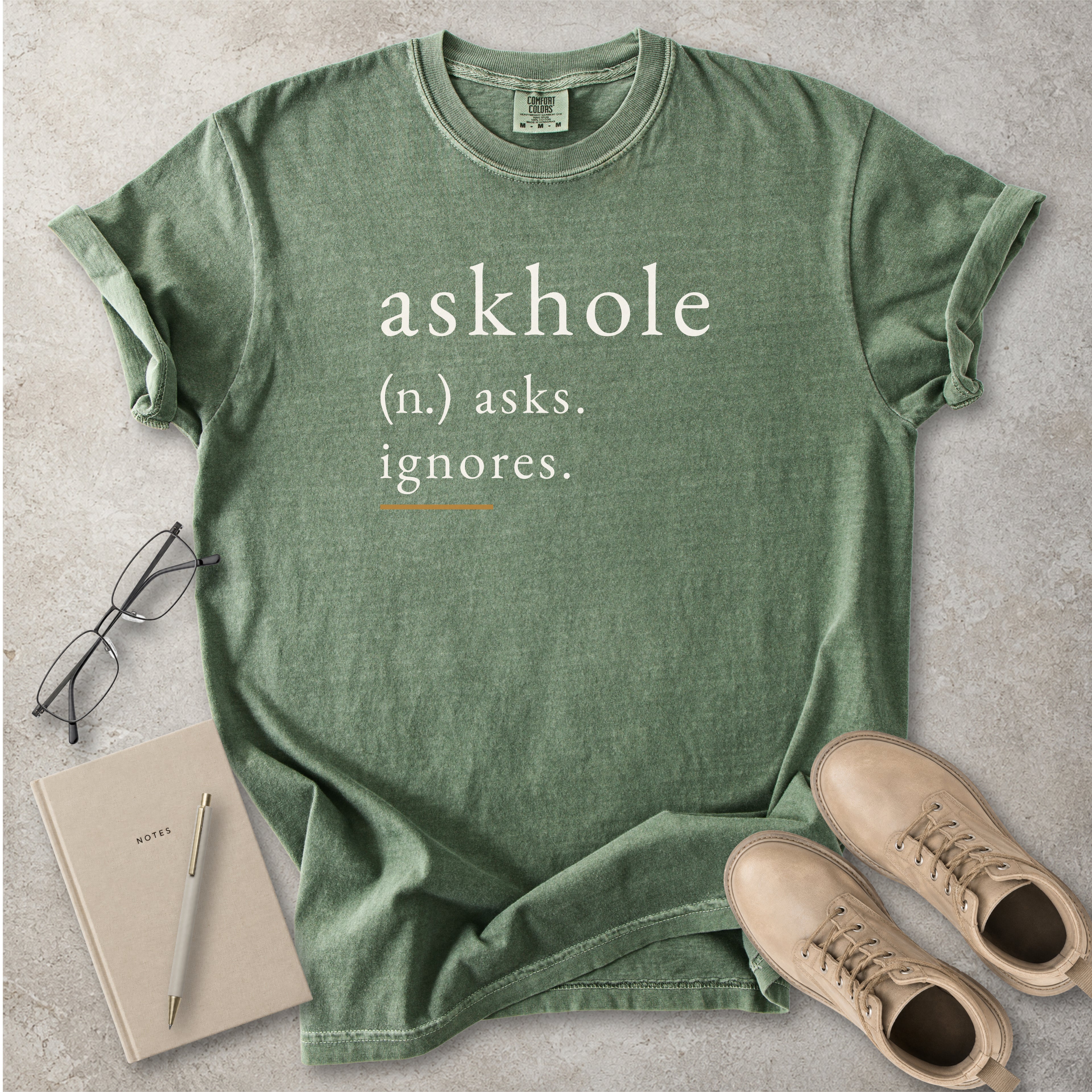 Moss green Comfort Colors graphic T-shirt with askhole definition text reading asks ignores, styled with notebook, pen, glasses and tan boots