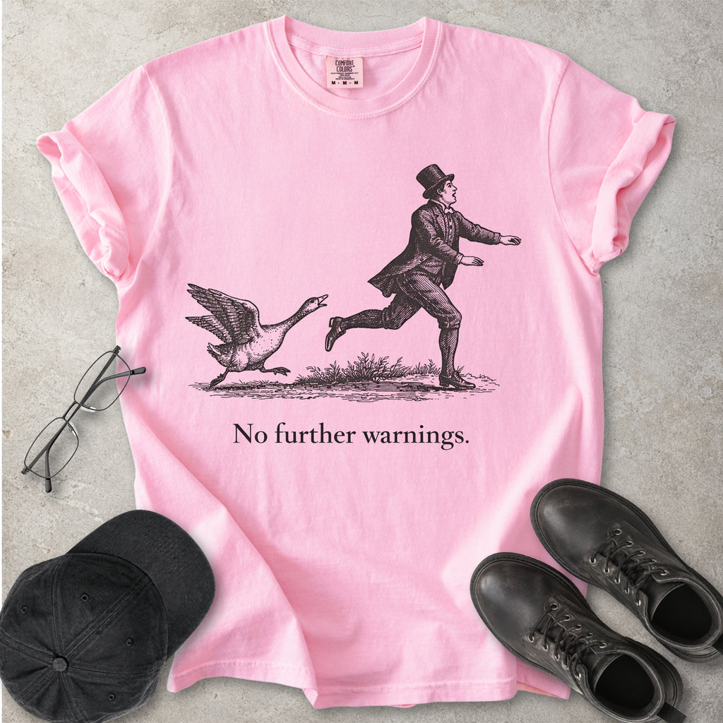 Pink t-shirt with a graphic of a man chasing a goose and text 'No further warnings.' on a concrete surface, surrounded by a cap, glasses, and boots.