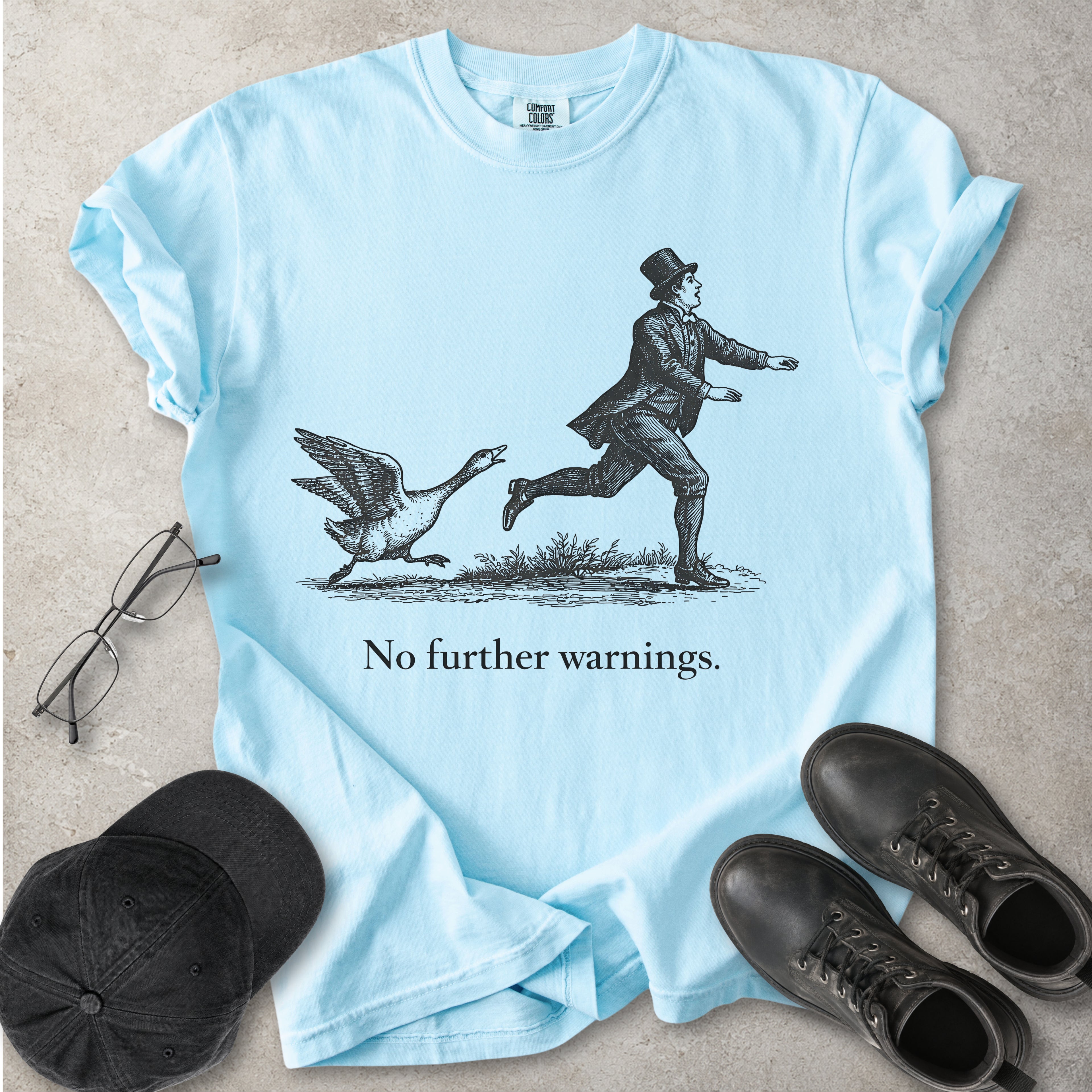 Light blue t-shirt with a graphic of a man running from a goose and text 'No further warnings.' on a concrete surface, surrounded by a cap, glasses, and boots.