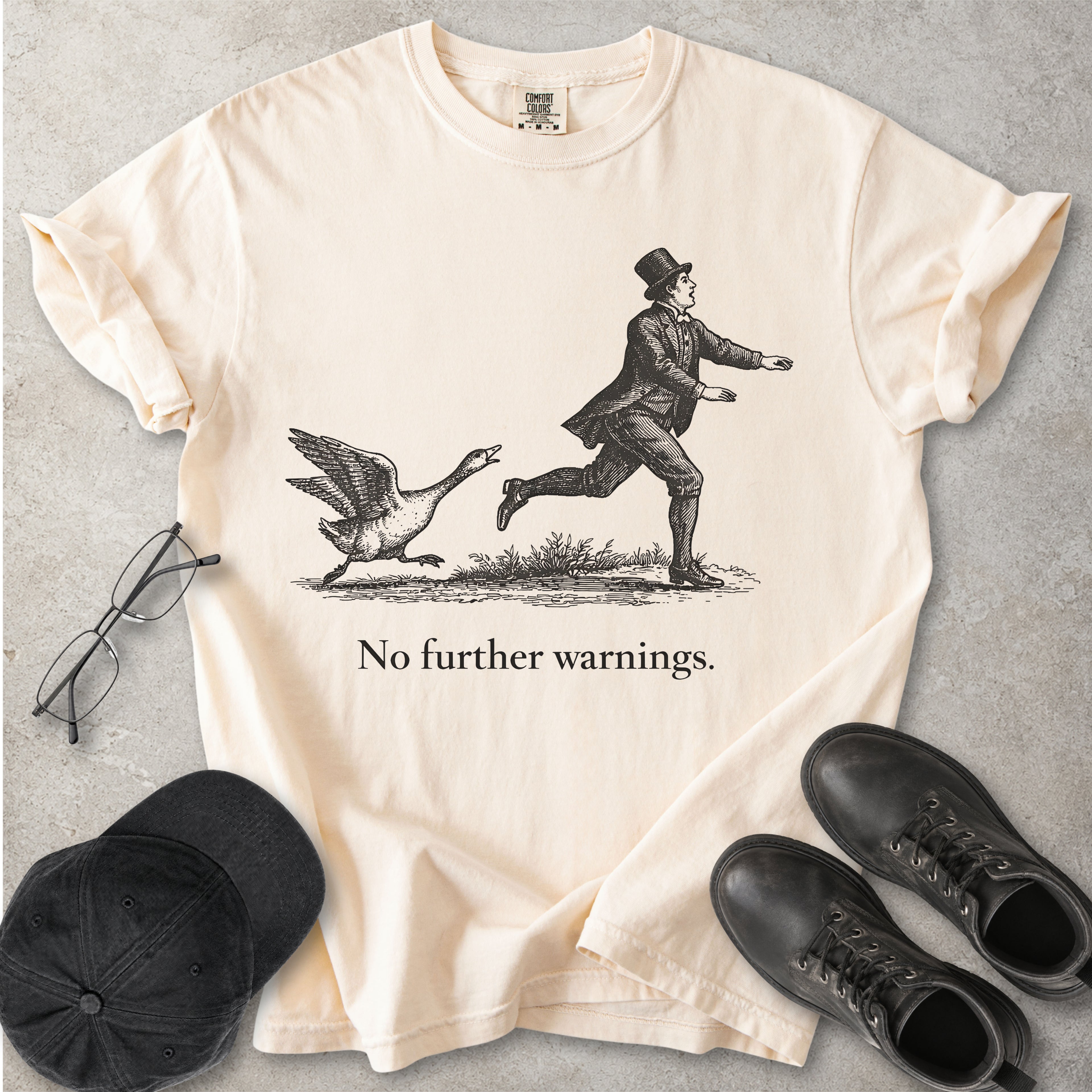 Ivory beige Comfort Colors graphic T-shirt with vintage black illustration of a goose chasing a man and text reading no further warnings, styled with black cap and boots