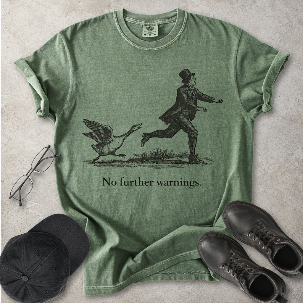 Green Comfort Colors graphic T-shirt with vintage black illustration of a goose chasing a man and text reading no further warnings, styled with black cap and boots