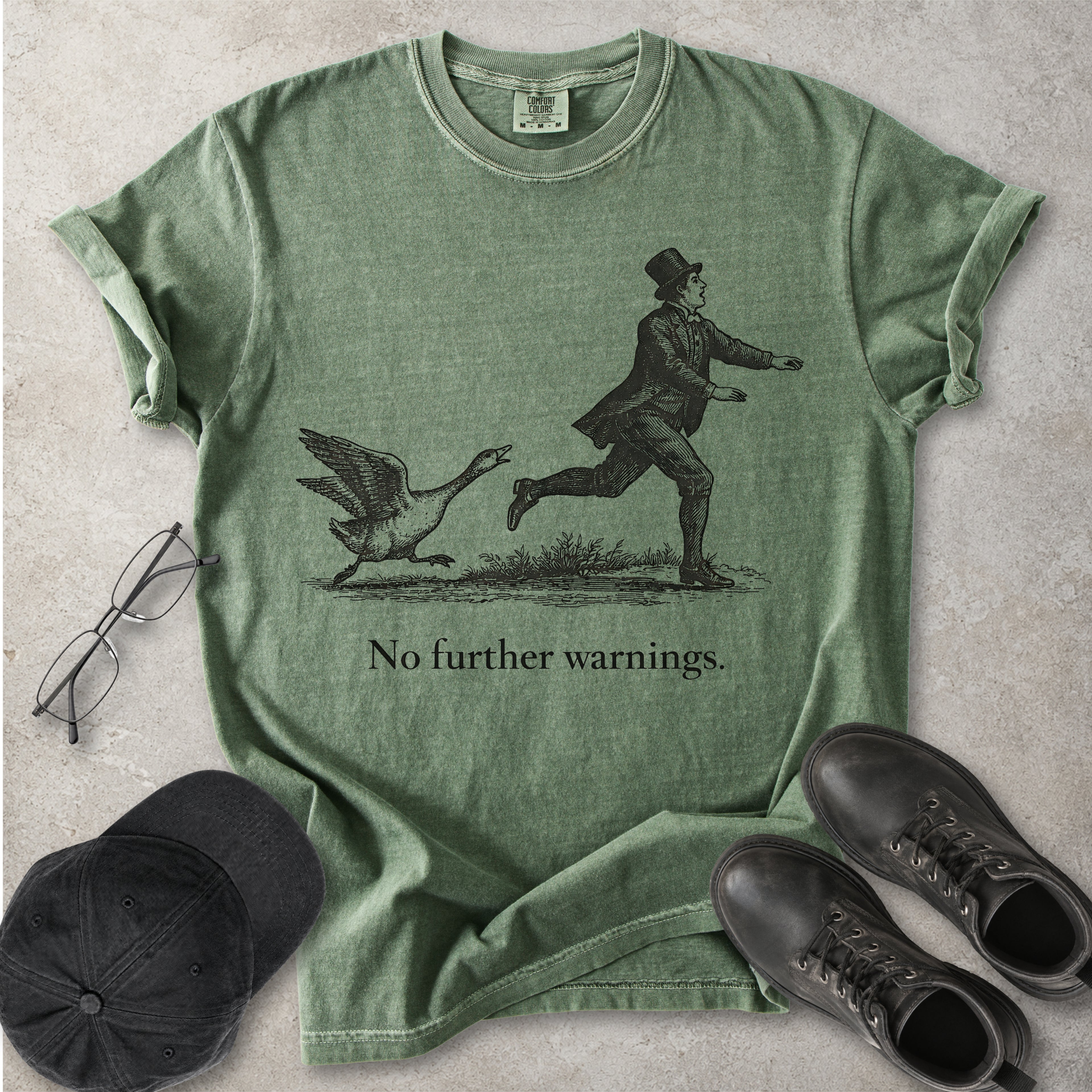 Green Comfort Colors graphic T-shirt with vintage black illustration of a goose chasing a man and text reading no further warnings, styled with black cap and boots
