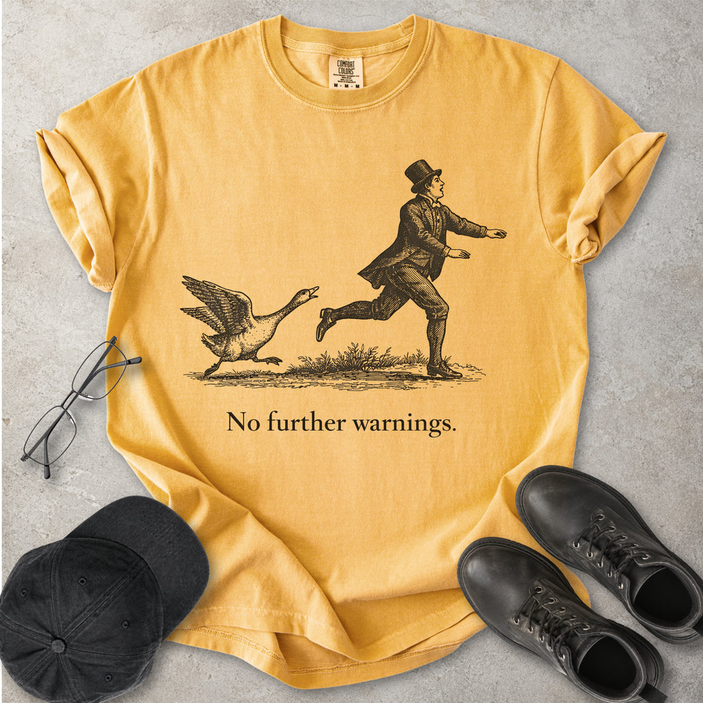 Yellow t-shirt with a graphic of a man running from a goose and text 'No further warnings.' on a gray surface with a cap and shoes.