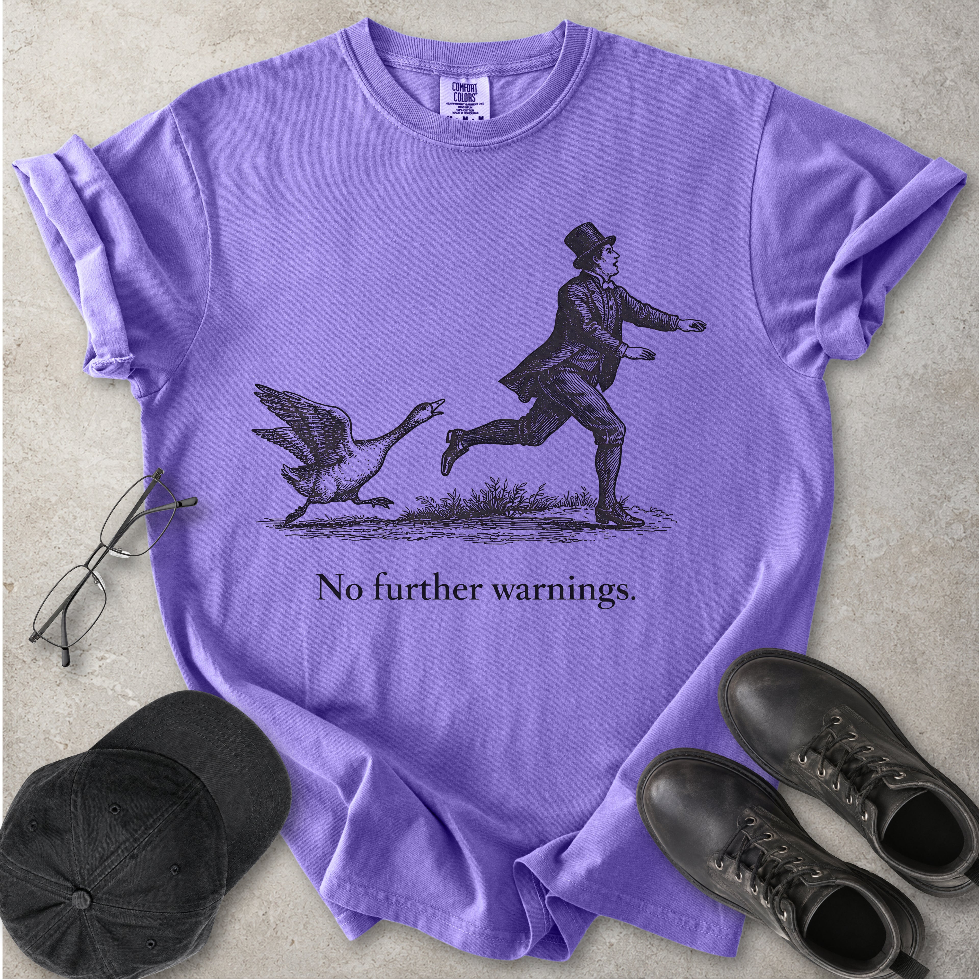 Violet purple Comfort Colors graphic T-shirt with vintage black illustration of a goose chasing a man and text reading no further warnings, styled with black cap and boots