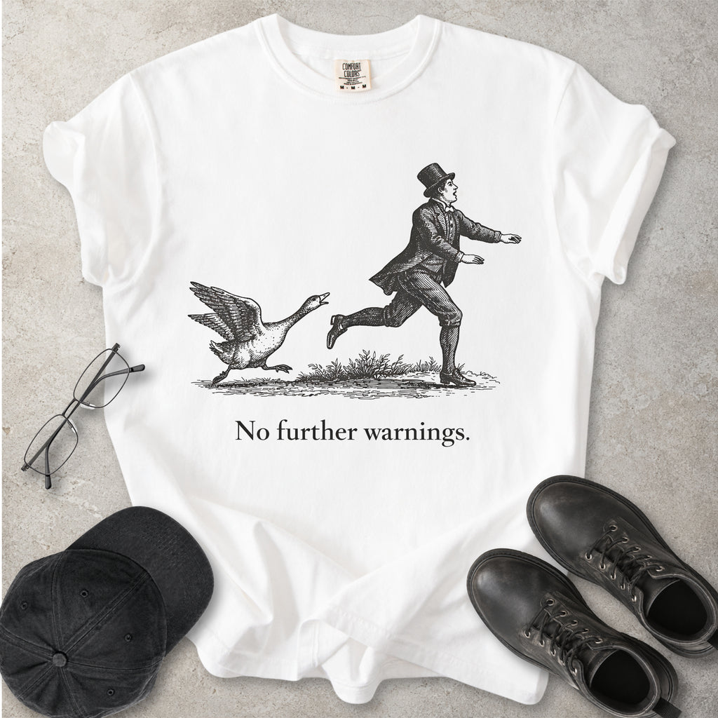 White t-shirt with a graphic of a man running from a goose and text 'No further warnings.' on a gray surface, surrounded by glasses, a cap, and shoes.