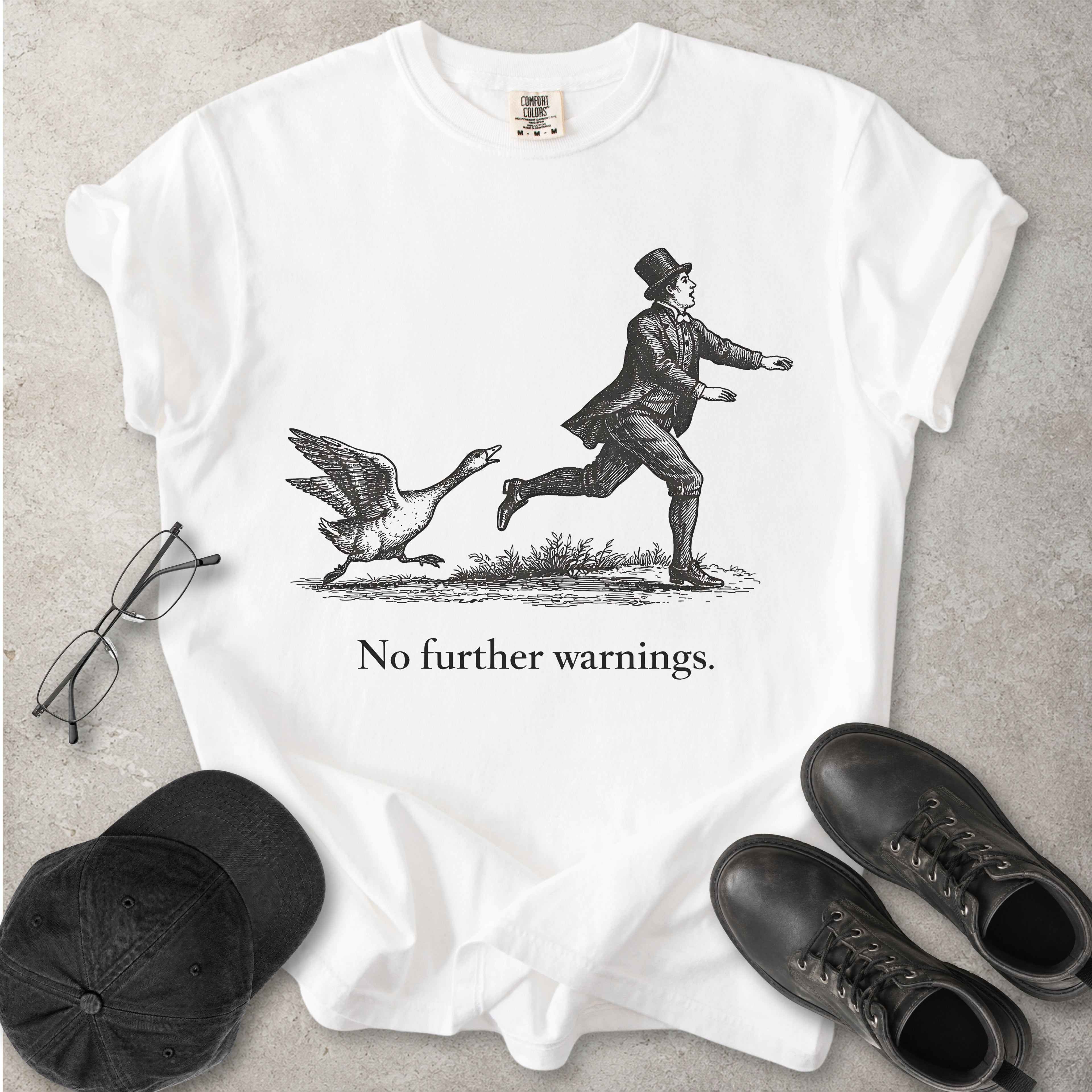 White t-shirt with a graphic of a man running from a goose and text 'No further warnings.' on a gray surface, surrounded by glasses, a cap, and shoes.