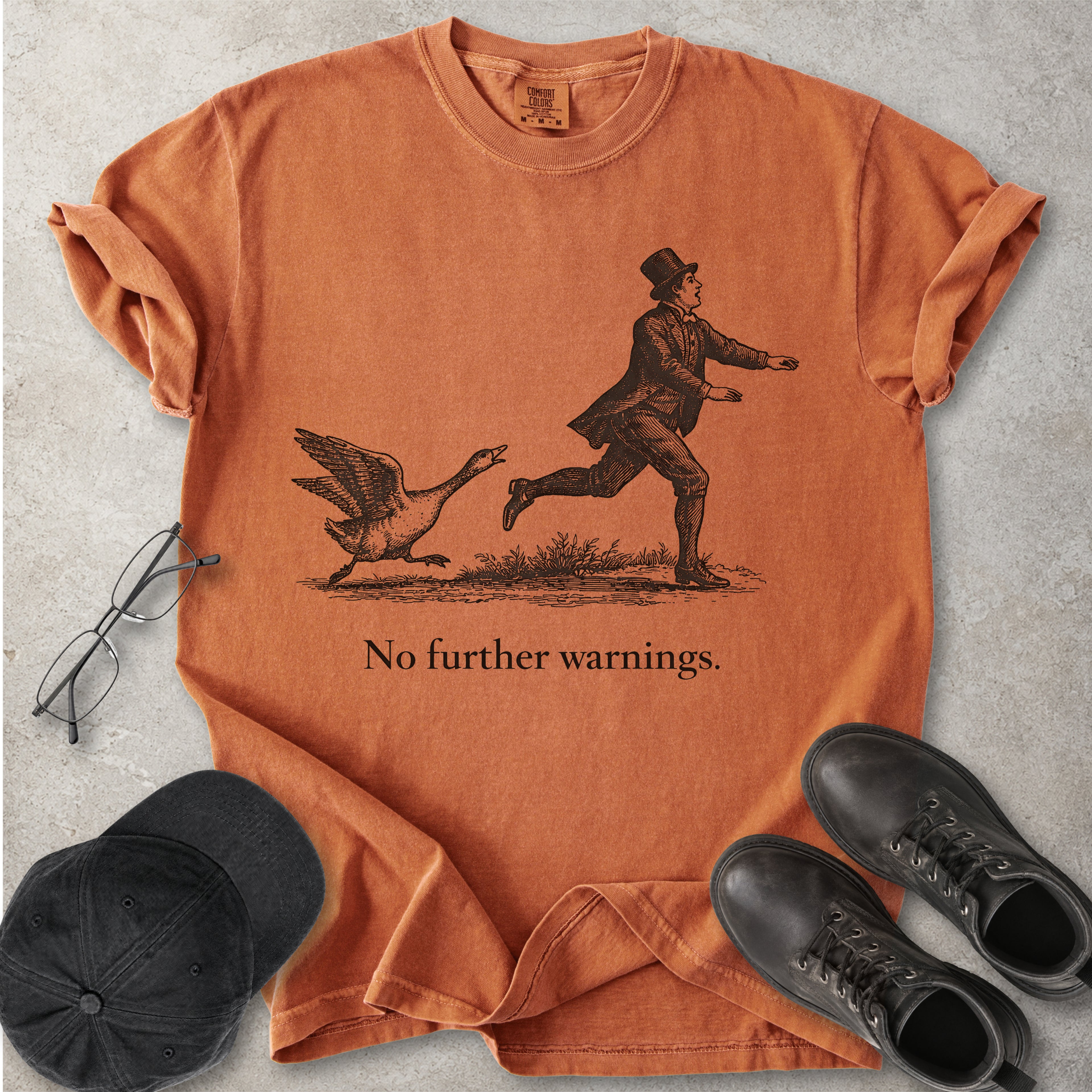 Yam orange Comfort Colors graphic T-shirt with vintage black illustration of a goose chasing a man and text reading no further warnings, styled with black cap and boots