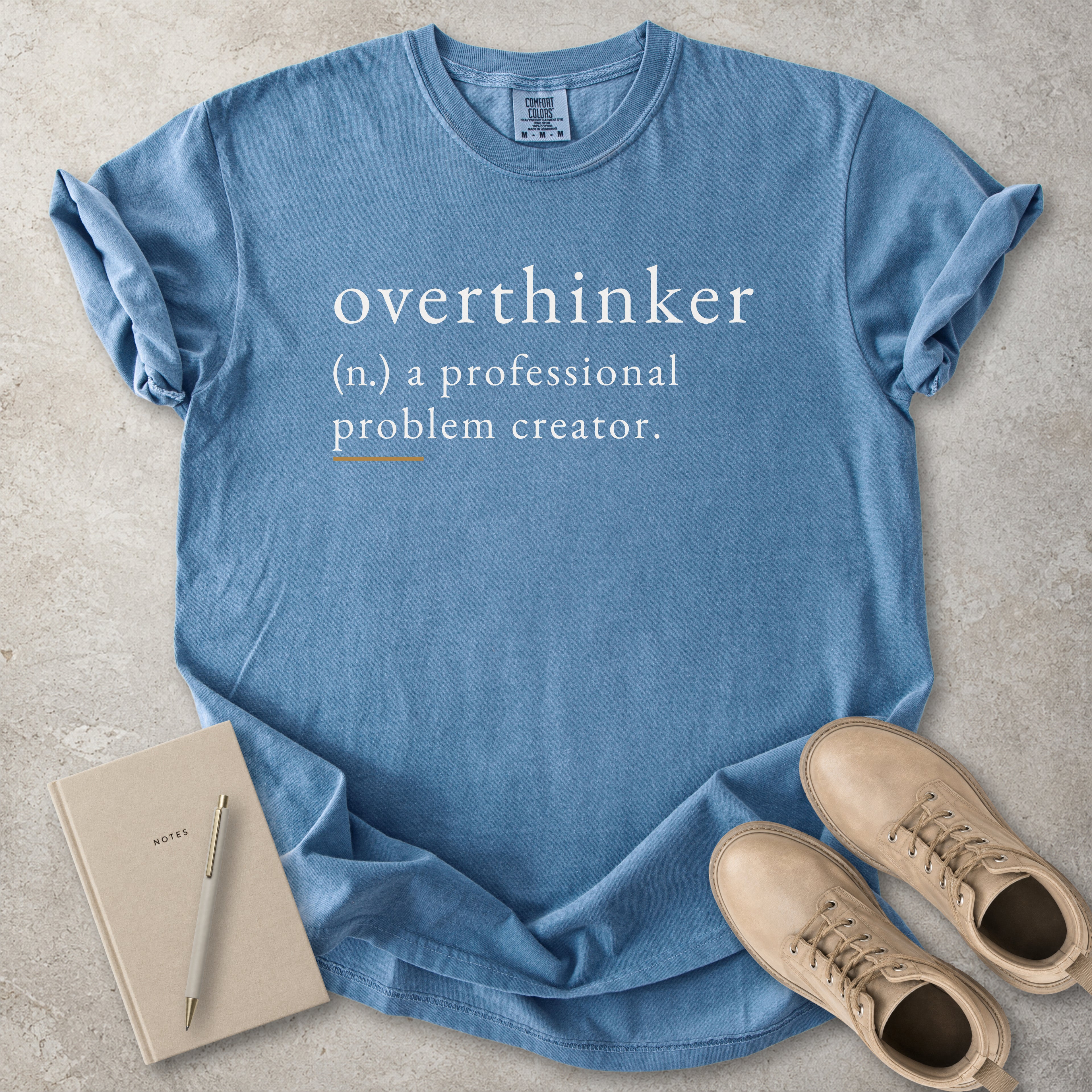 Blue Comfort Colors graphic T-shirt with overthinker definition text reading a professional problem creator, styled with notebook, pen and tan boots