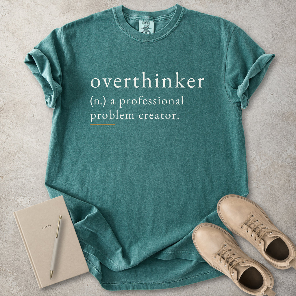 Green Comfort Colors graphic T-shirt with overthinker definition text reading a professional problem creator, styled with notebook, pen and tan boots