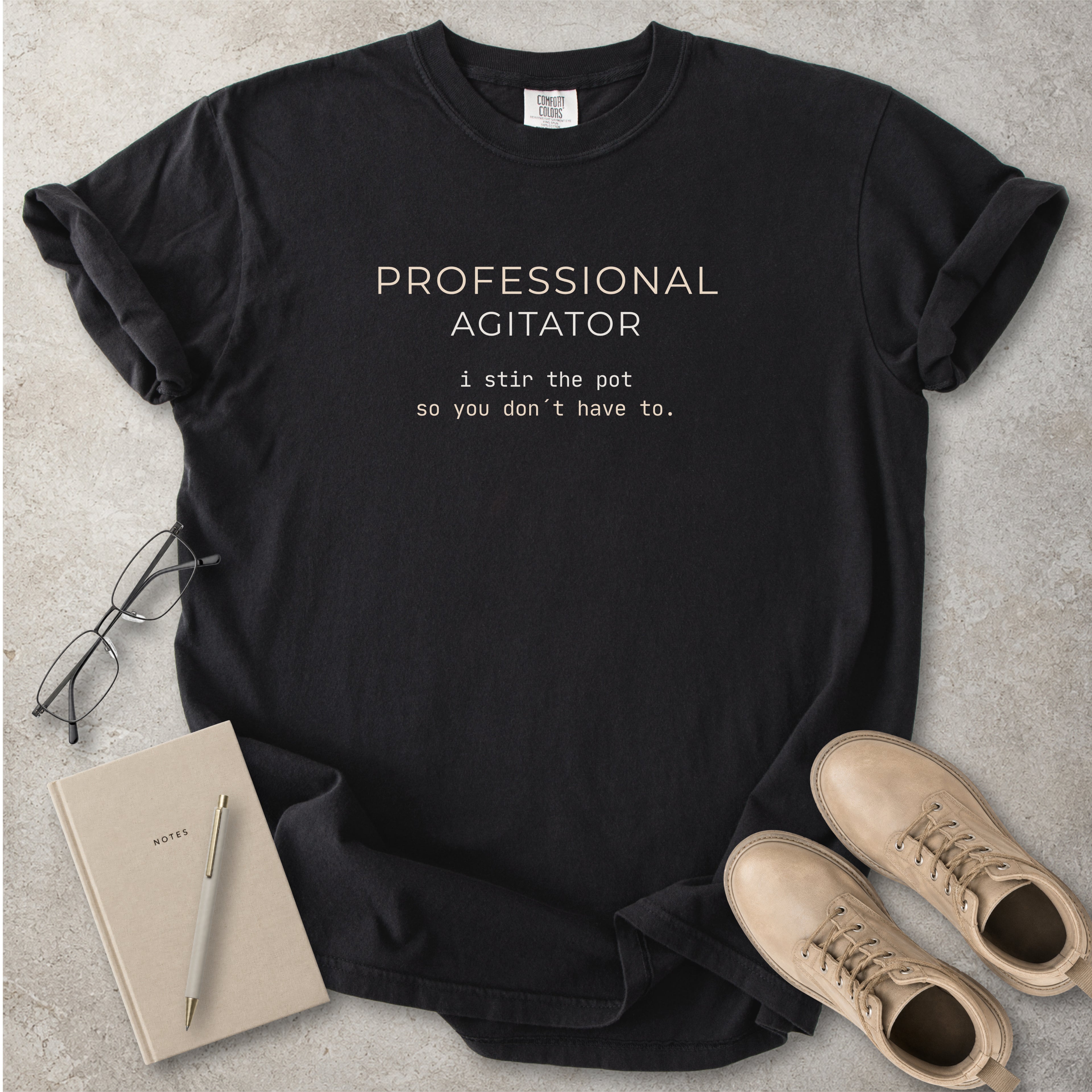 Black Comfort Colors graphic T-shirt with beige text reading Professional Agitator, I stir the pot so you don’t have to, styled with notebook, pen and tan boots
