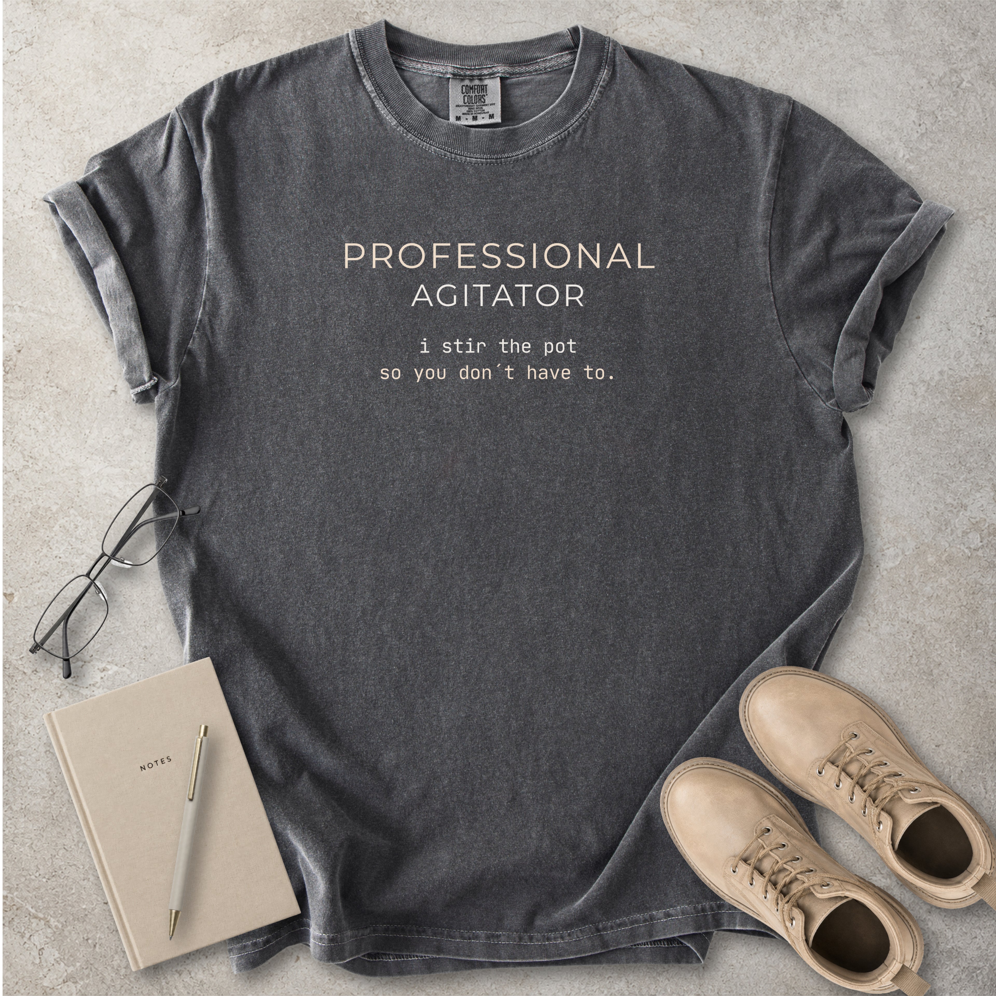 Grey Comfort Colors graphic T-shirt with beige text reading Professional Agitator, I stir the pot so you don’t have to, styled with notebook, pen and tan boots