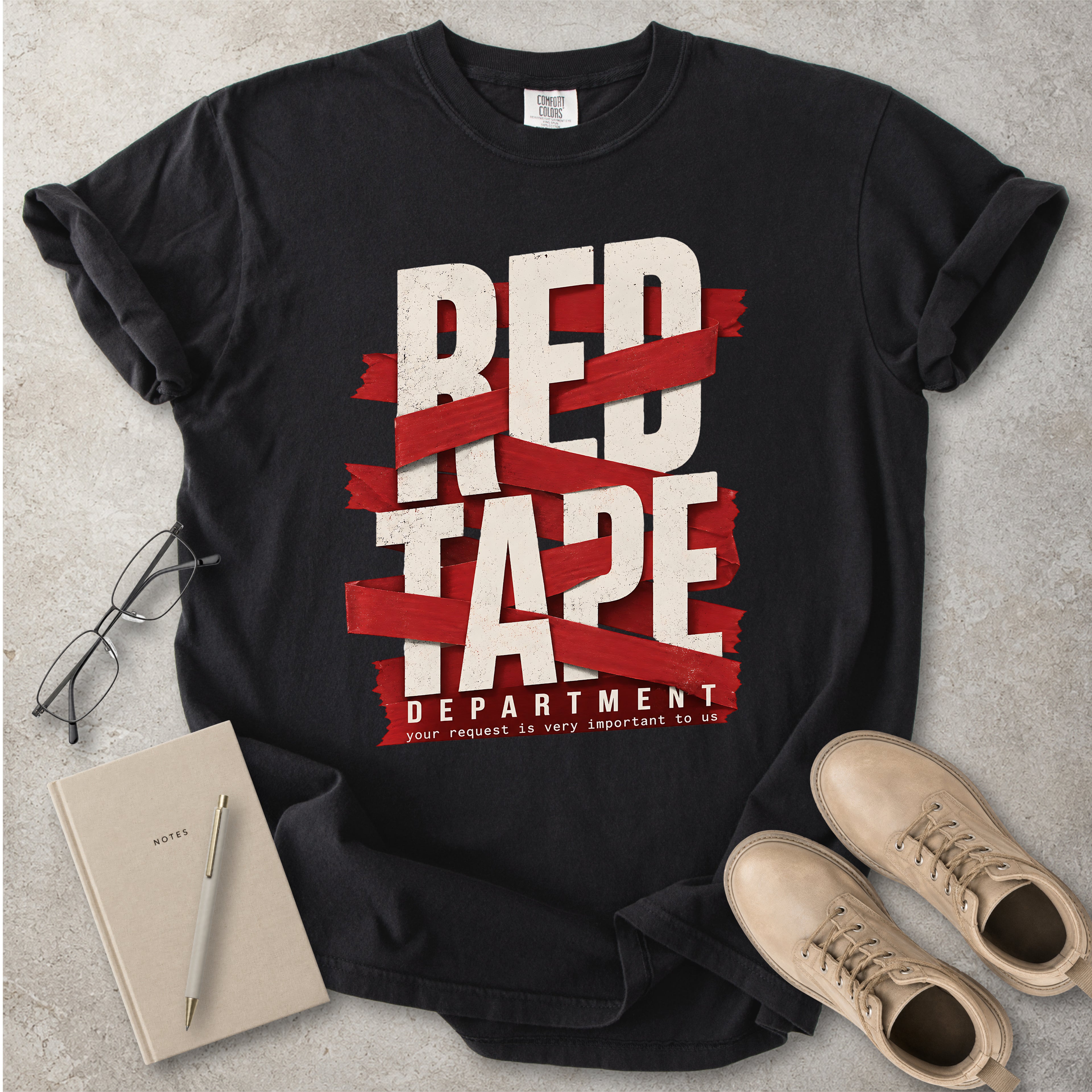 Black t-shirt with 'Red Tape Department' graphic on a gray surface with notebook and boots.