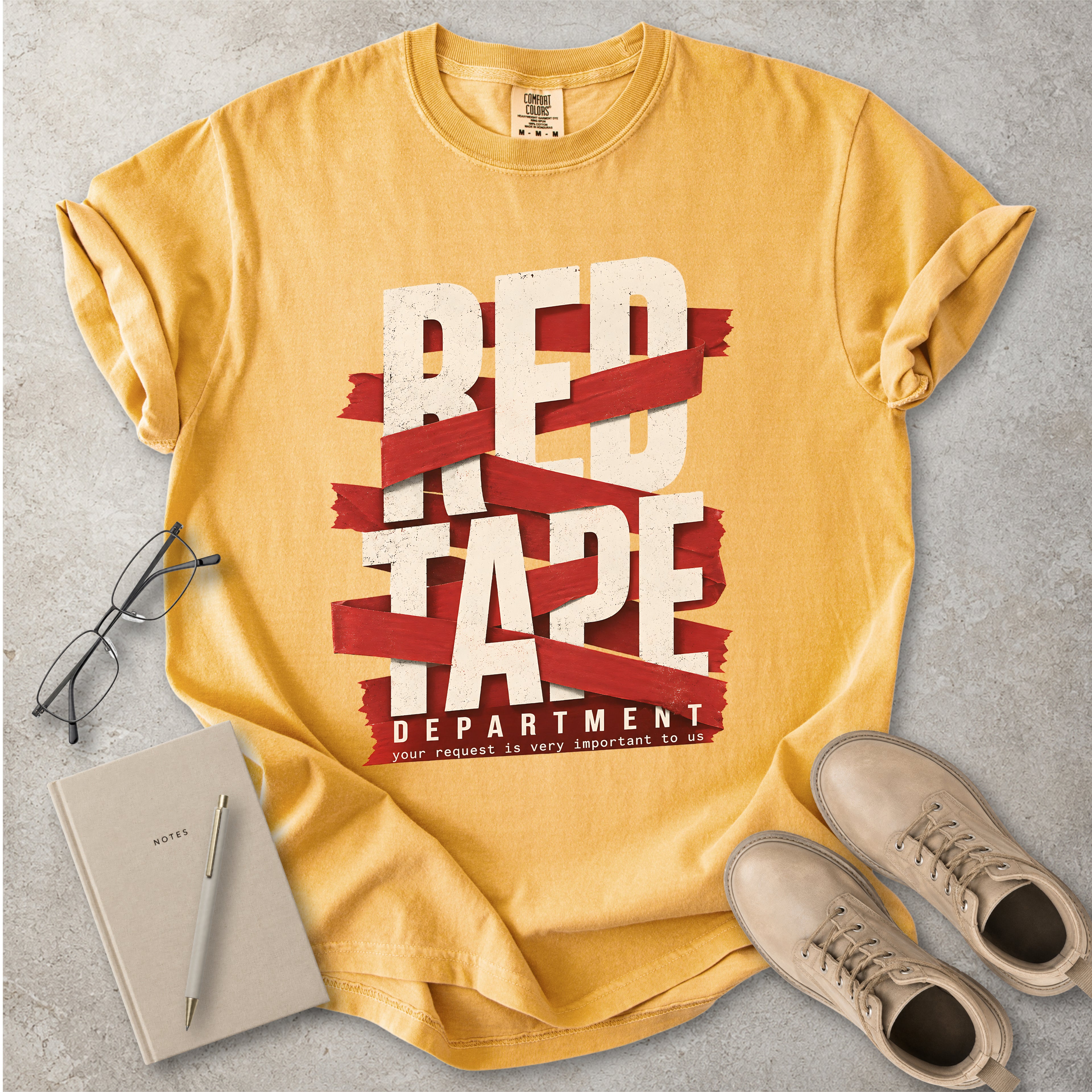 Yellow t-shirt with 'Red Tape Department' design on a gray surface with glasses, notebook, and shoes.