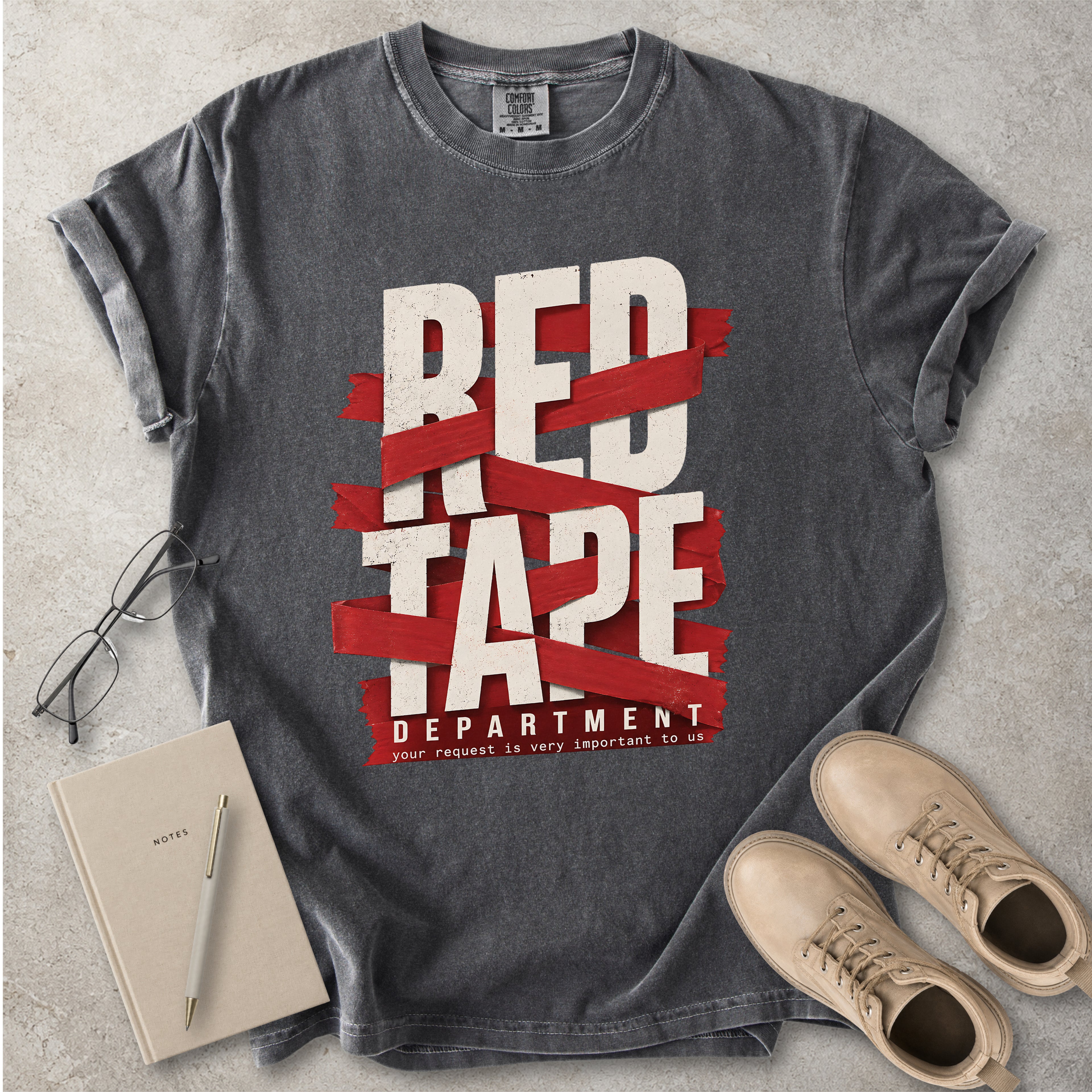 Gray t-shirt with 'Red Tape Department' design on a concrete surface with notebook, pen, and boots.