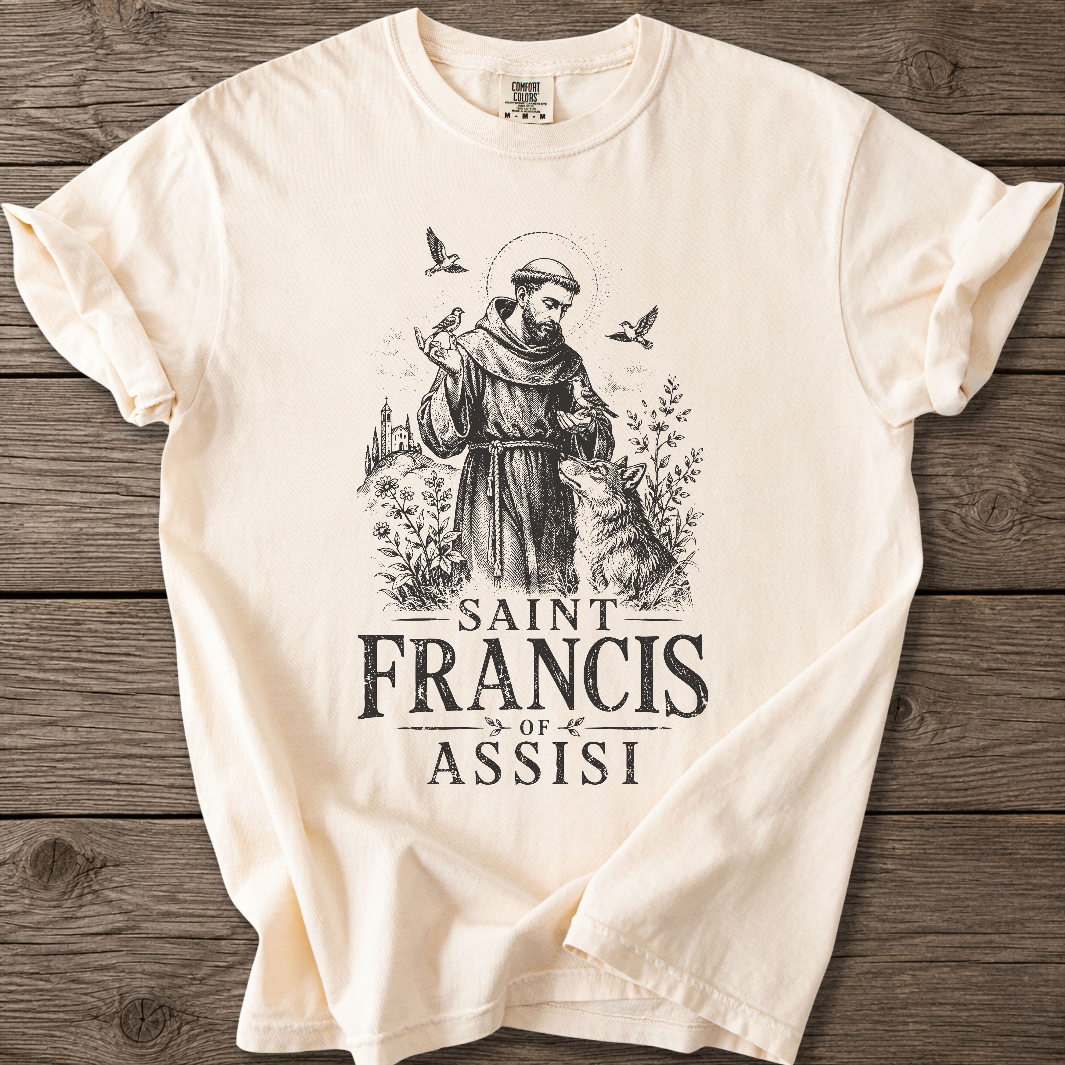 An off-white short-sleeve t-shirt with a black graphic of Saint Francis of Assisi, displayed flat on a dark wood background.