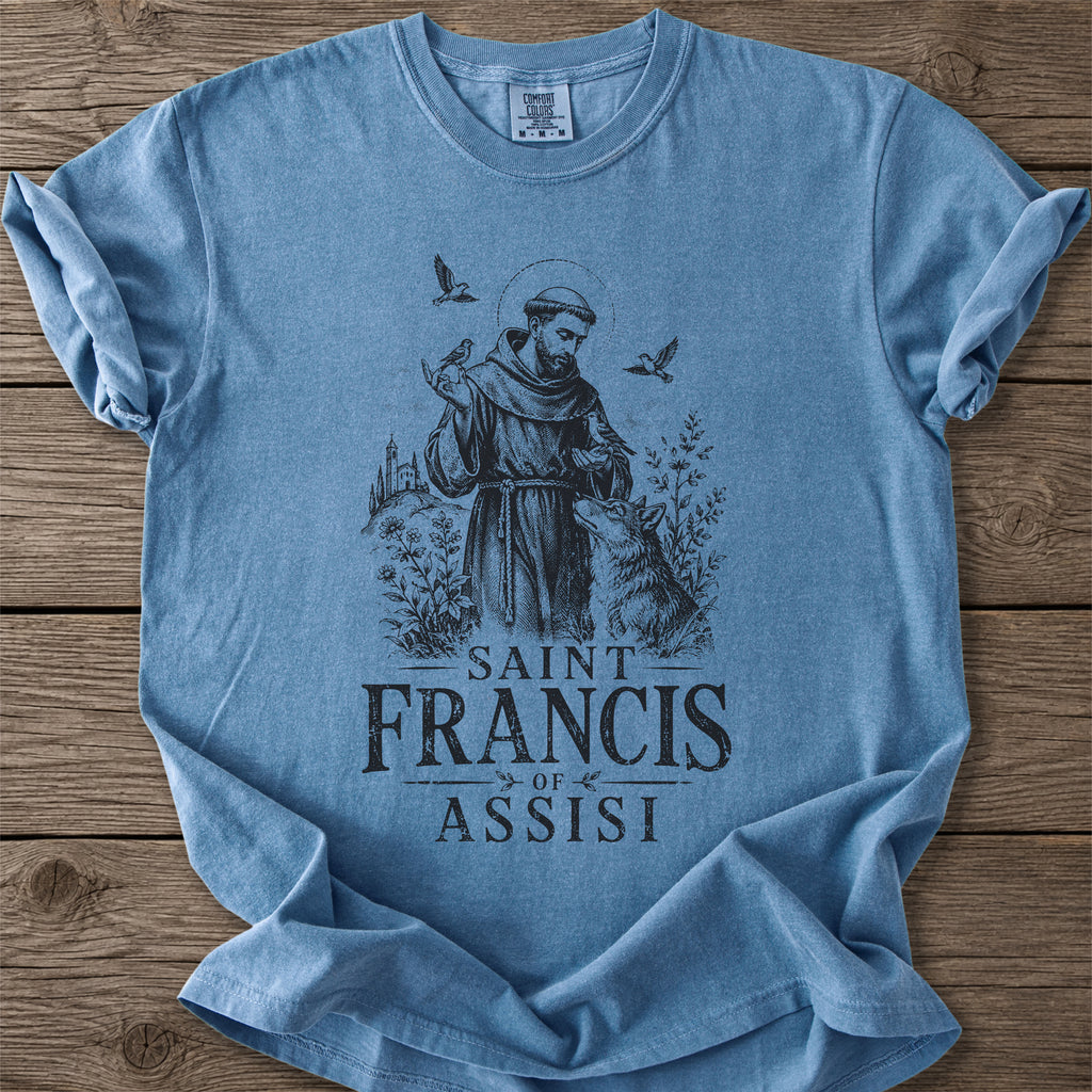 A light blue crewneck t-shirt featuring a dark blue graphic of Saint Francis of Assisi, laid flat on a rustic wooden background.