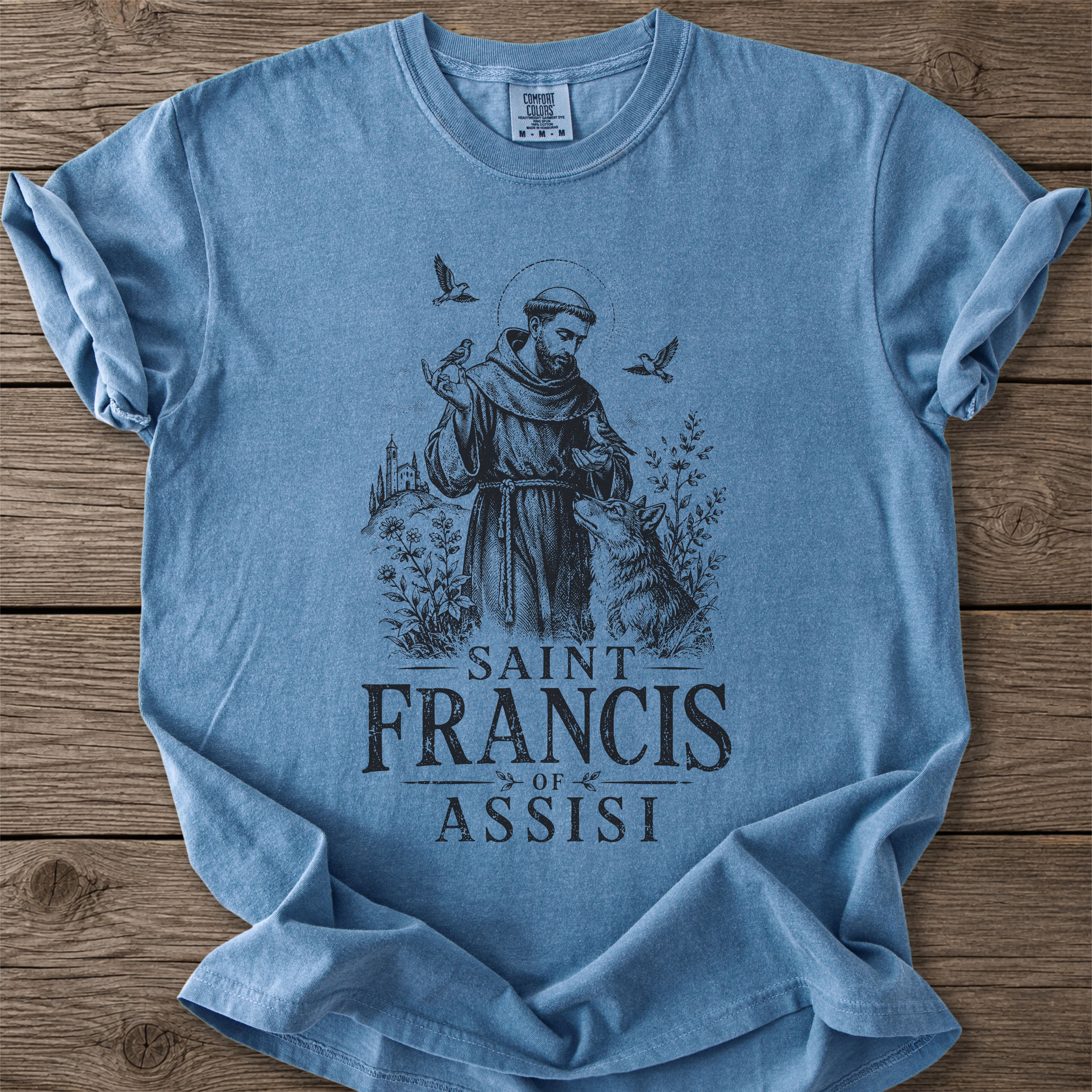 A light blue crewneck t-shirt featuring a dark blue graphic of Saint Francis of Assisi, laid flat on a rustic wooden background.