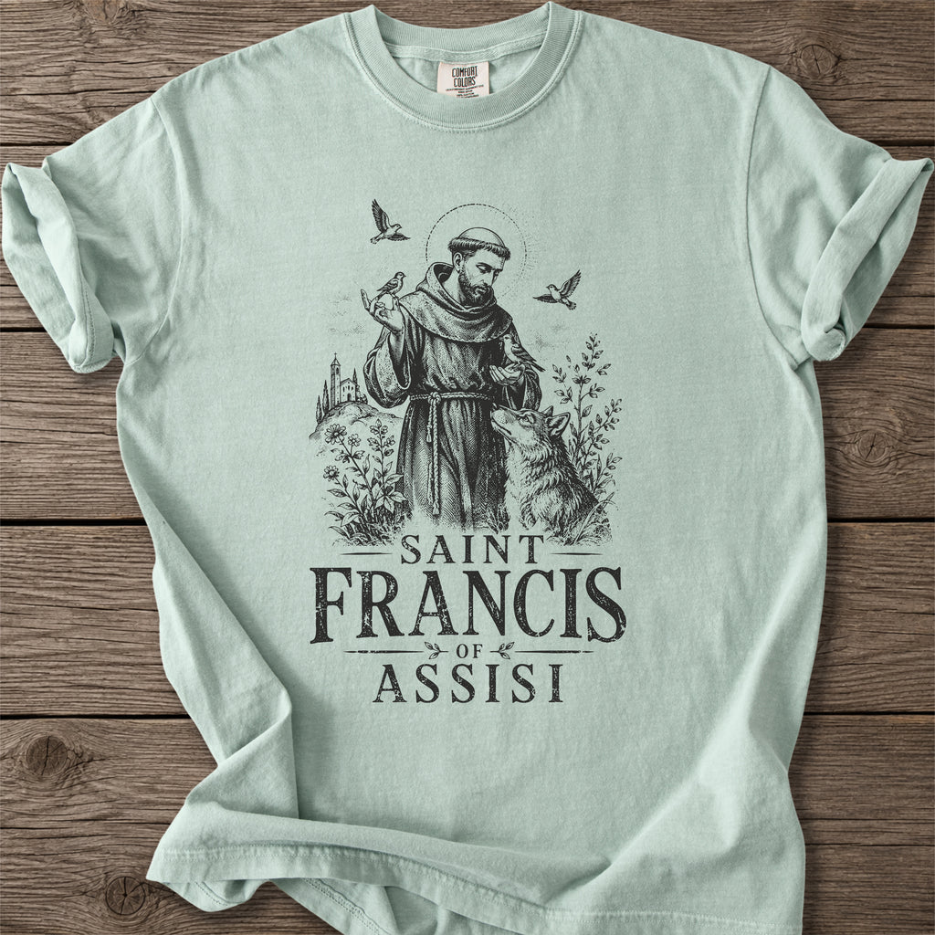 A light mint green t-shirt with a black graphic of Saint Francis of Assisi, displayed flat on a rustic wooden background.