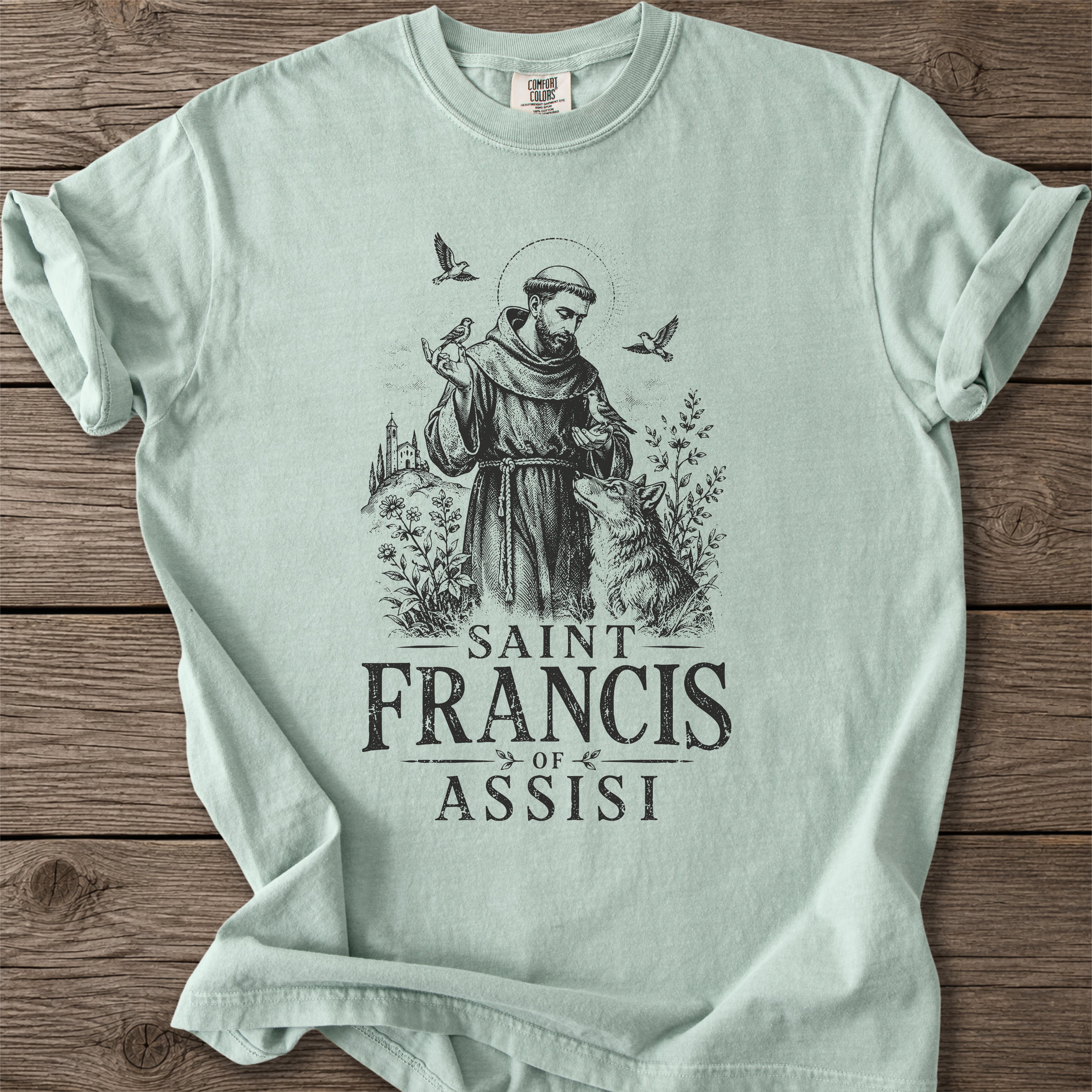 A light mint green t-shirt with a black graphic of Saint Francis of Assisi, displayed flat on a rustic wooden background.