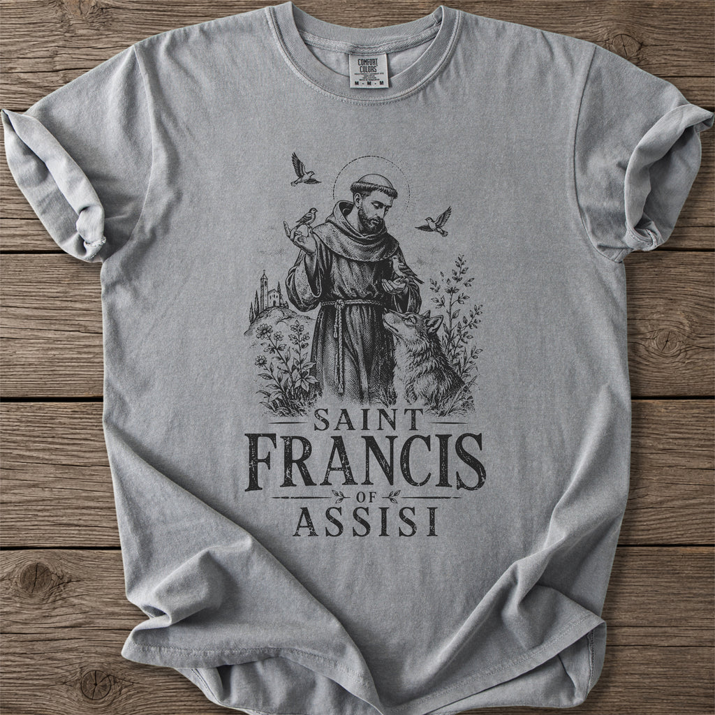 A light gray crewneck t-shirt featuring a black graphic print of Saint Francis of Assisi with a rooster and the text 'SAINT FRANCIS OF ASSISI' underneath, laid flat on a rustic wooden background.