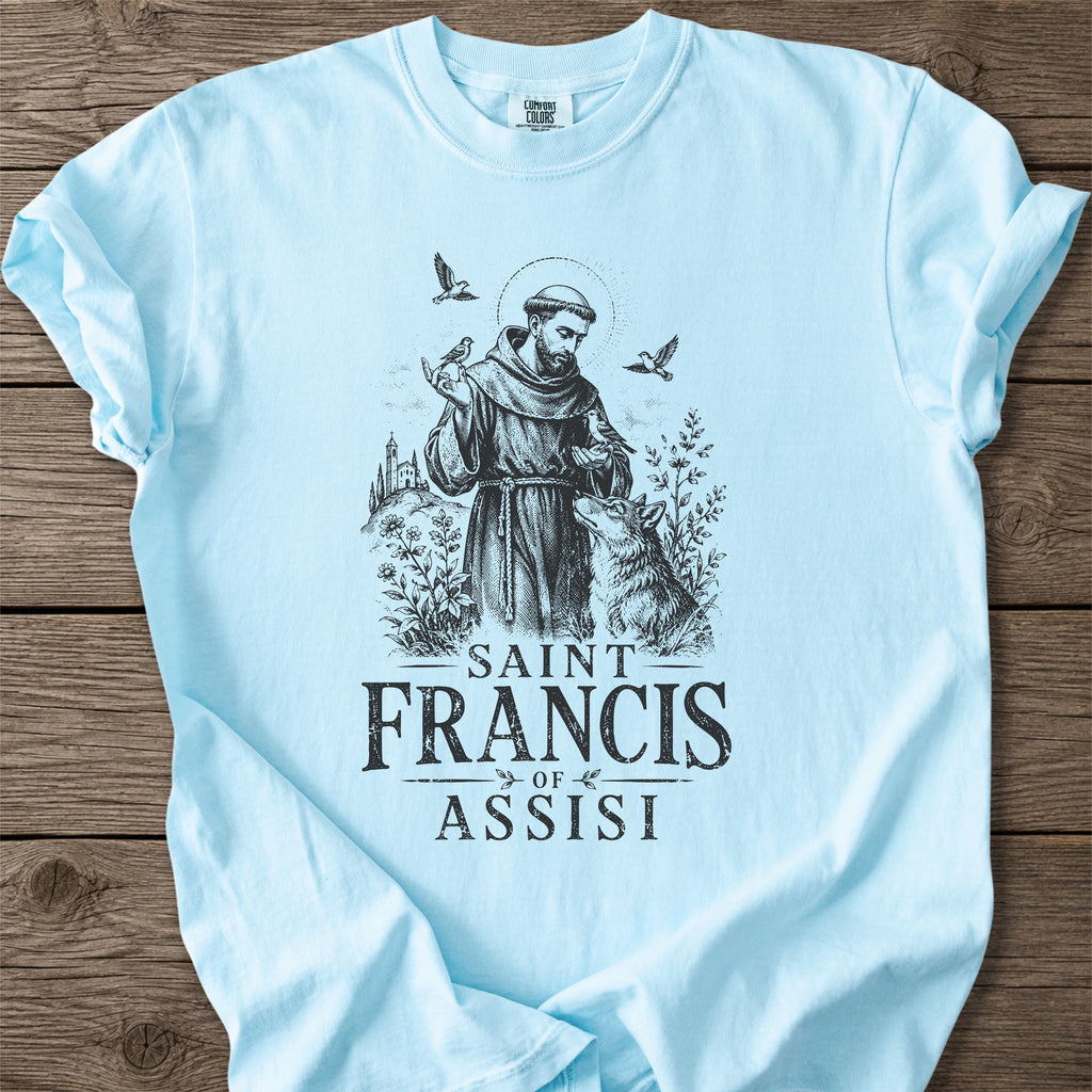 A light blue short-sleeve t-shirt laid flat on a rustic wooden background, featuring a black graphic print of Saint Francis of Assisi with a wolf and the text 'SAINT FRANCIS OF ASSISI'.
