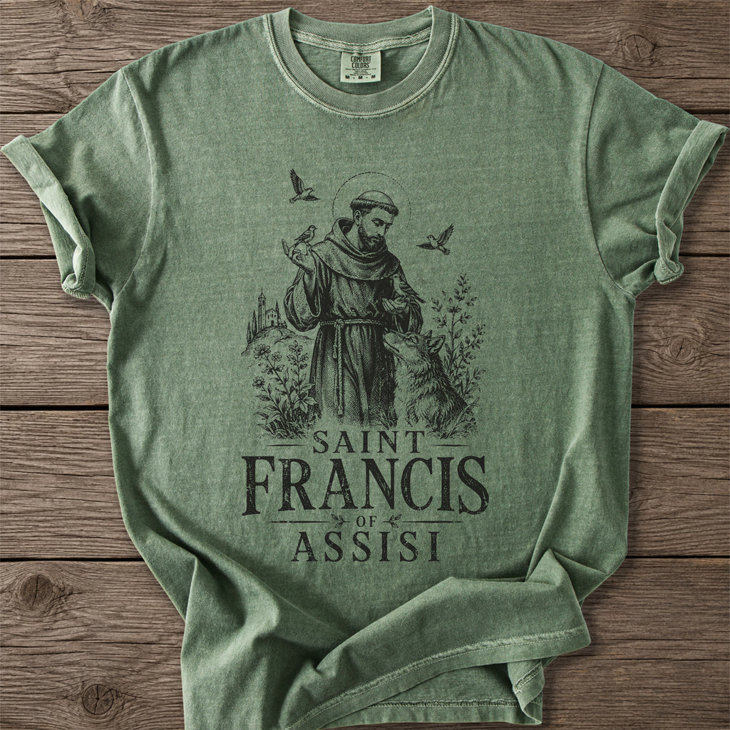 A sage green heathered t-shirt featuring a dark graphic of Saint Francis of Assisi with a wolf, birds, and the text 'SAINT FRANCIS OF ASSISI' printed on the front, laid flat on a rustic wooden background.