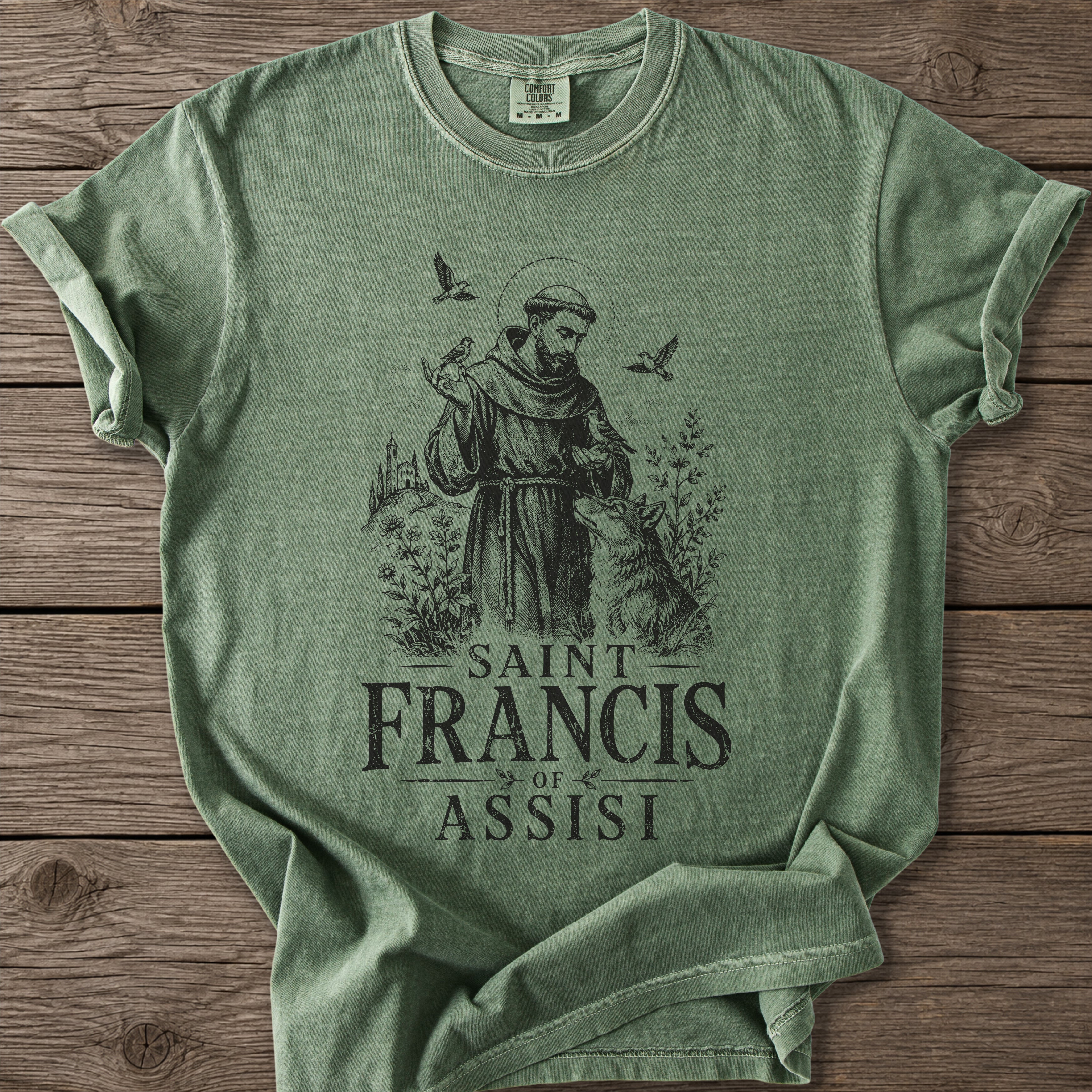 A sage green heathered t-shirt featuring a dark graphic of Saint Francis of Assisi with a wolf, birds, and the text 'SAINT FRANCIS OF ASSISI' printed on the front, laid flat on a rustic wooden background.