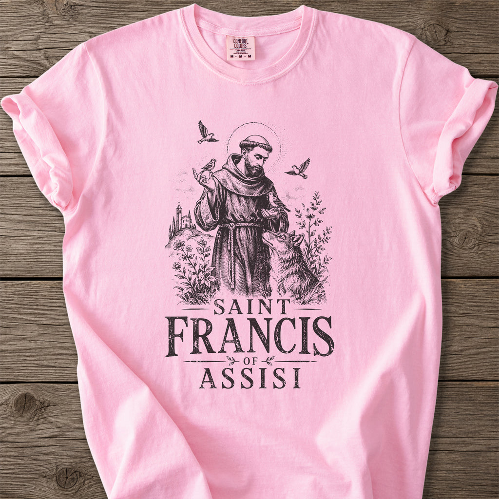 A light pink short-sleeve t-shirt with a black graphic print of Saint Francis of Assisi and the text 'SAINT FRANCIS OF ASSISI' laid flat on a rustic wooden background.