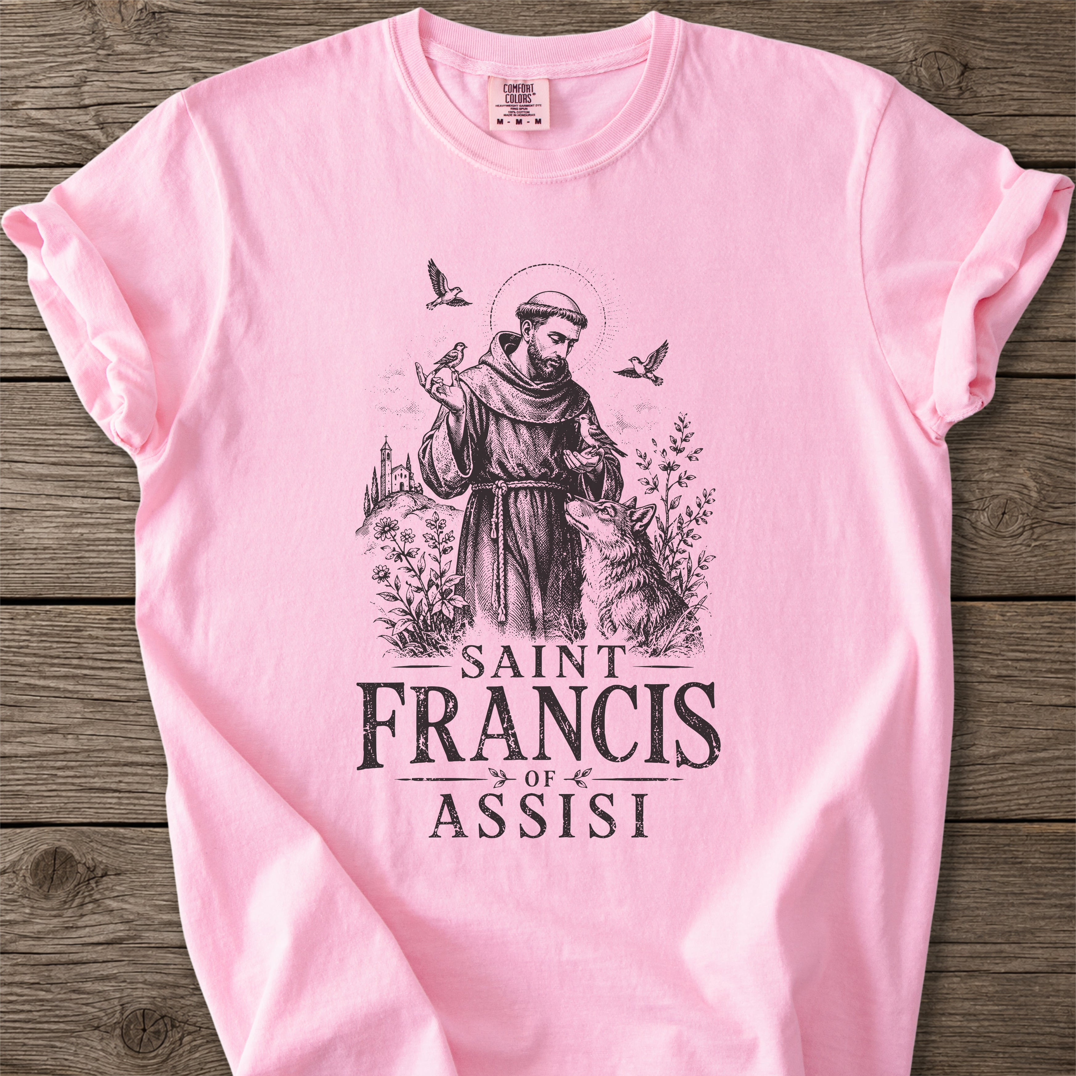 A light pink short-sleeve t-shirt with a black graphic print of Saint Francis of Assisi and the text 'SAINT FRANCIS OF ASSISI' laid flat on a rustic wooden background.
