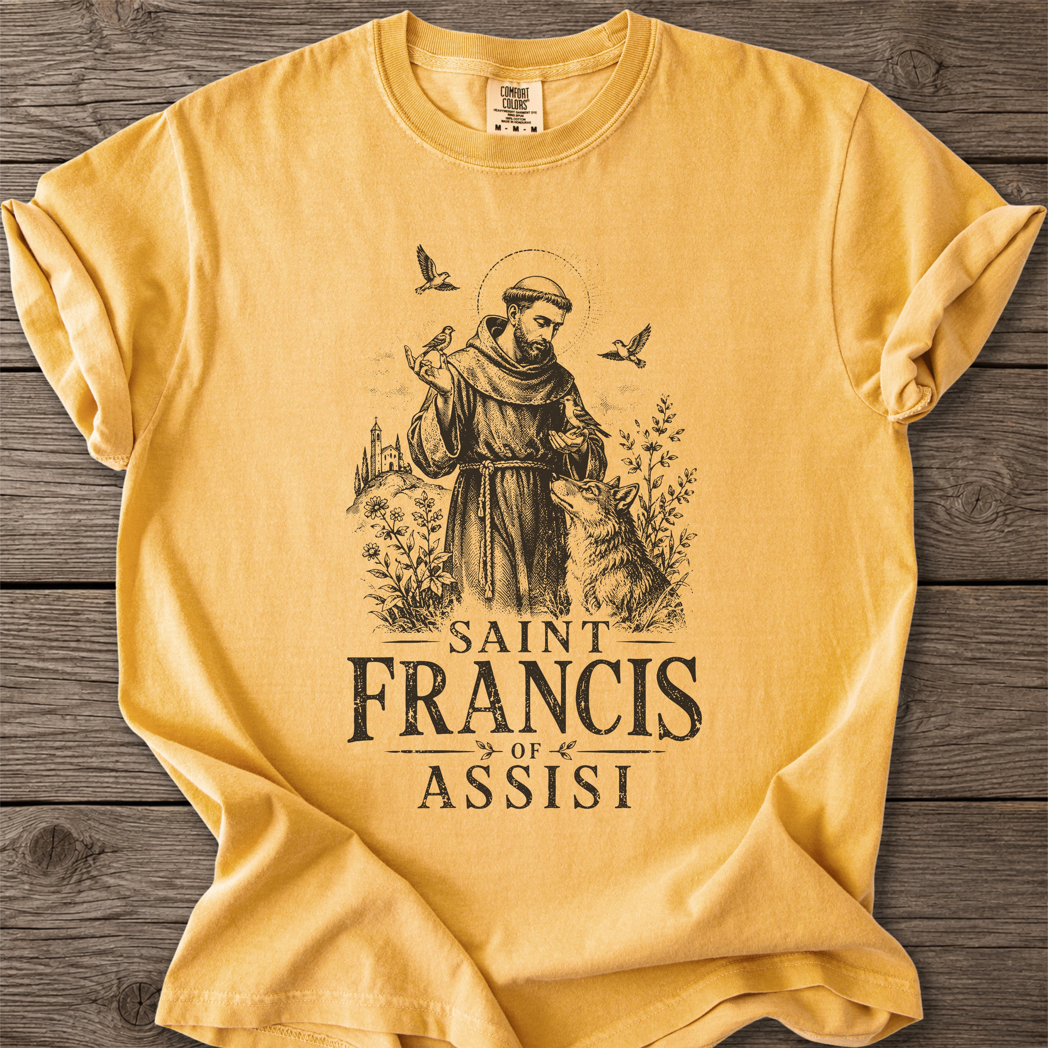 A mustard yellow short-sleeve t-shirt with a black graphic of Saint Francis of Assisi, displayed flat on a dark wood plank background.