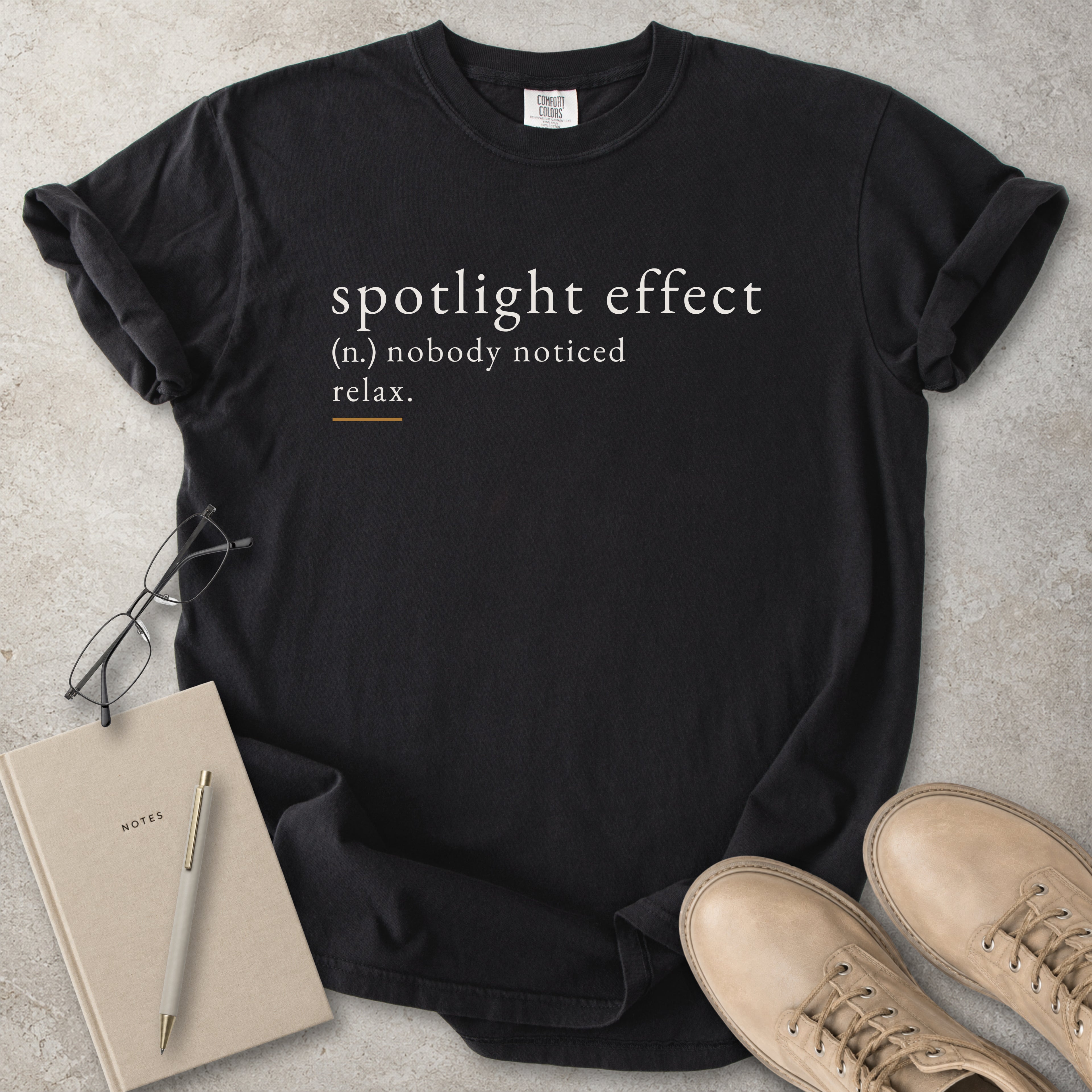 Black t-shirt with 'spotlight effect' text on a concrete surface with glasses, notebook, and shoes.
