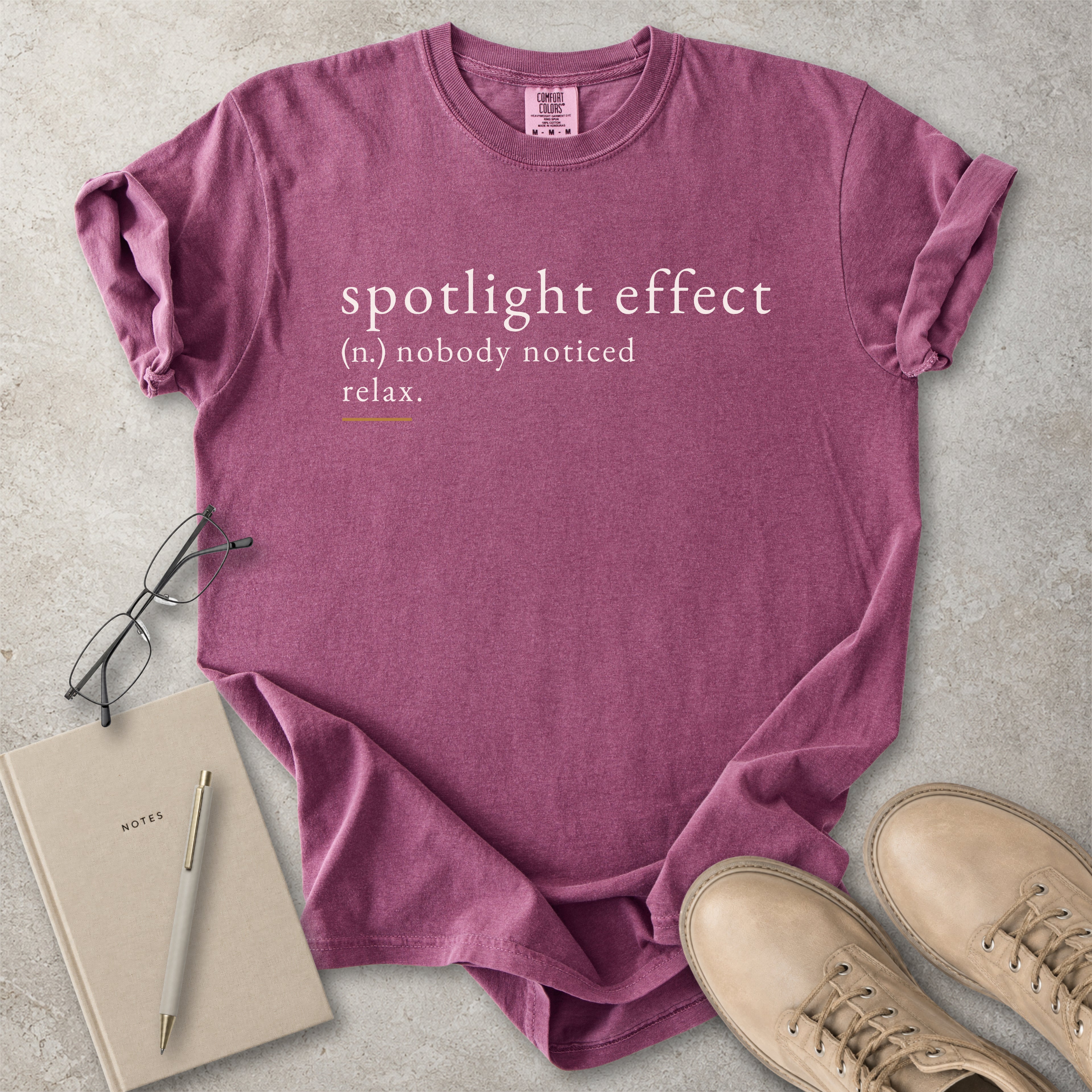 Purple t-shirt with 'spotlight effect' text on a concrete surface with notebook, pen, and shoes.