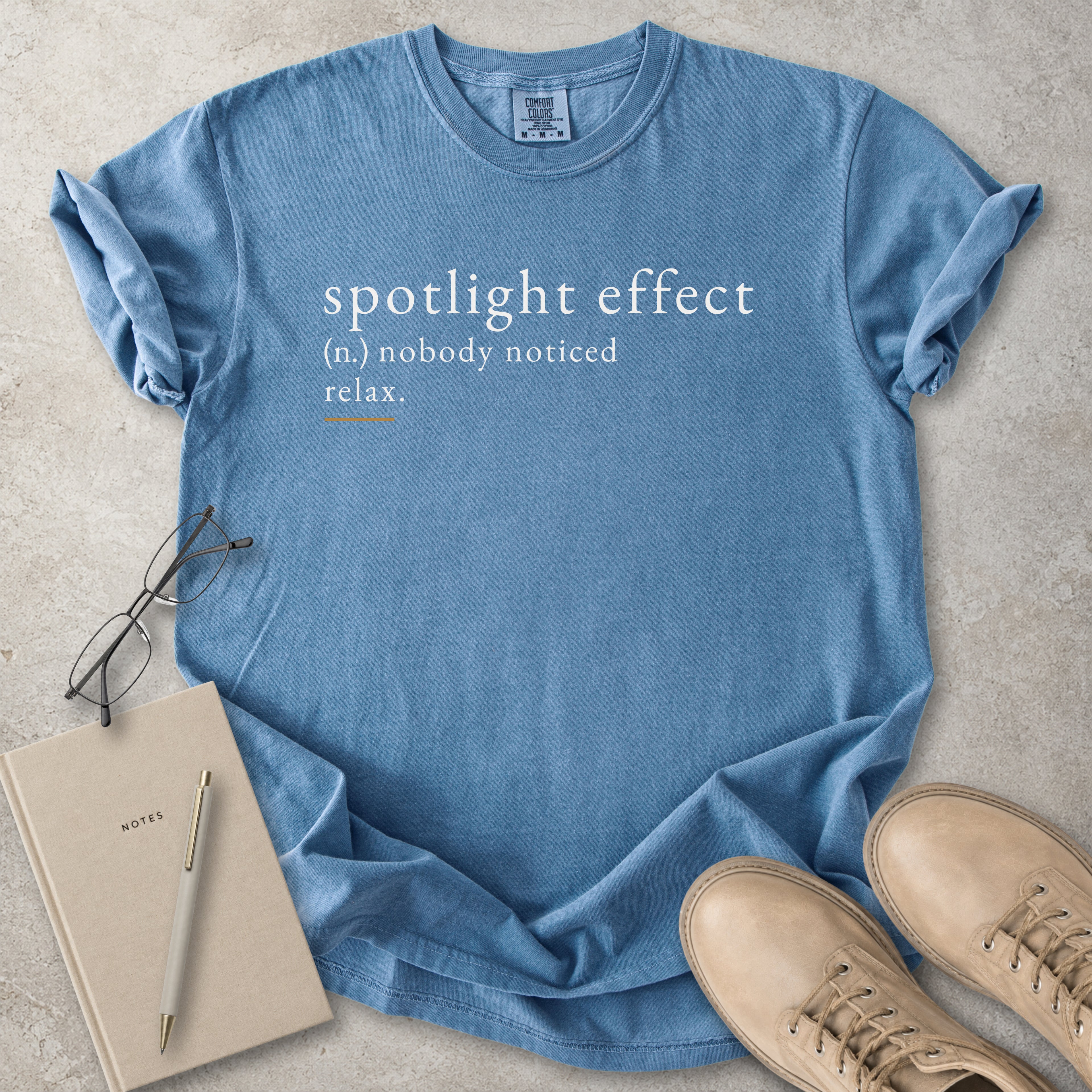 Blue Comfort Colors graphic T-shirt with spotlight effect definition text reading nobody noticed relax, styled with notebook, pen and tan boots