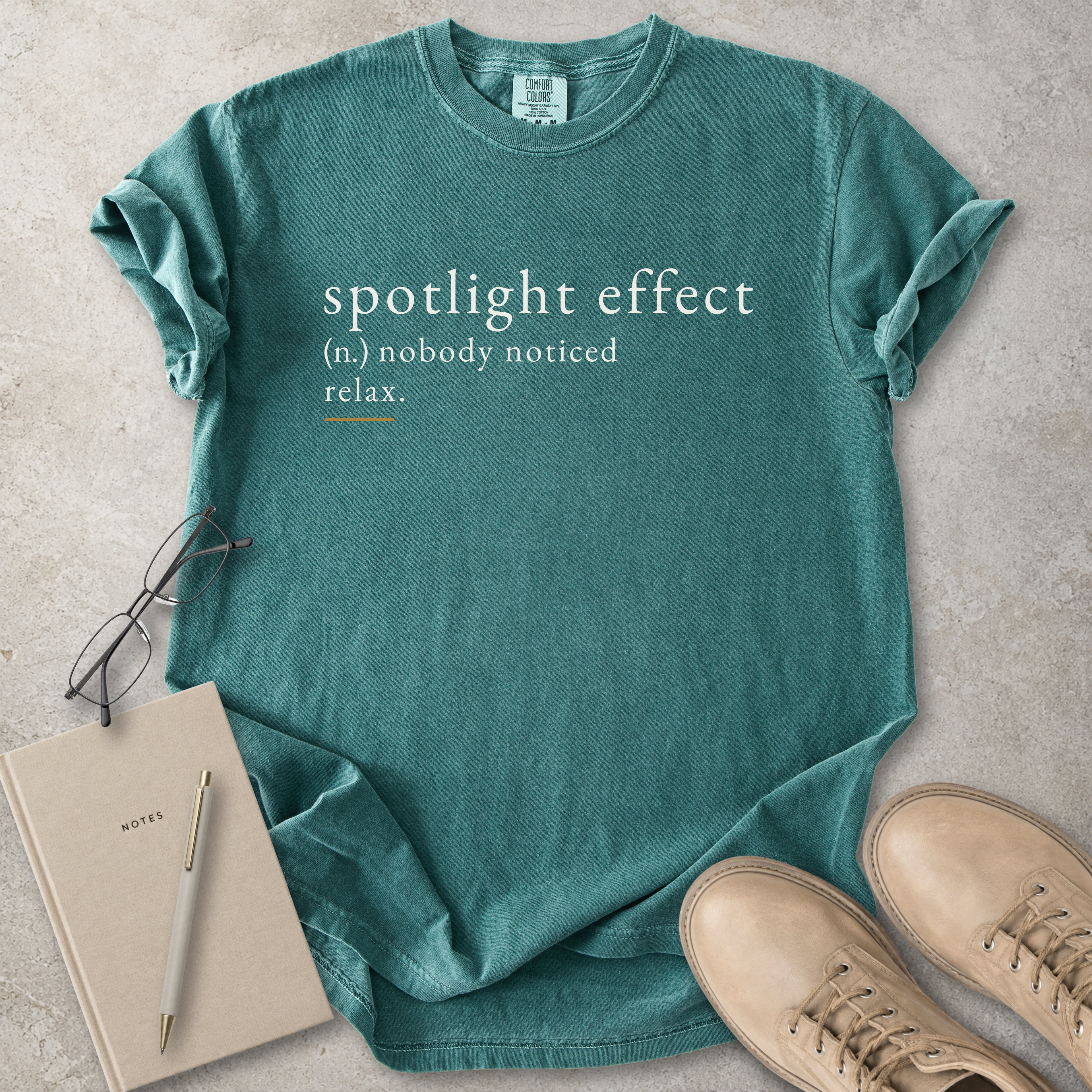 Teal t-shirt with 'spotlight effect' text, notebook, pen, and shoes on a concrete surface