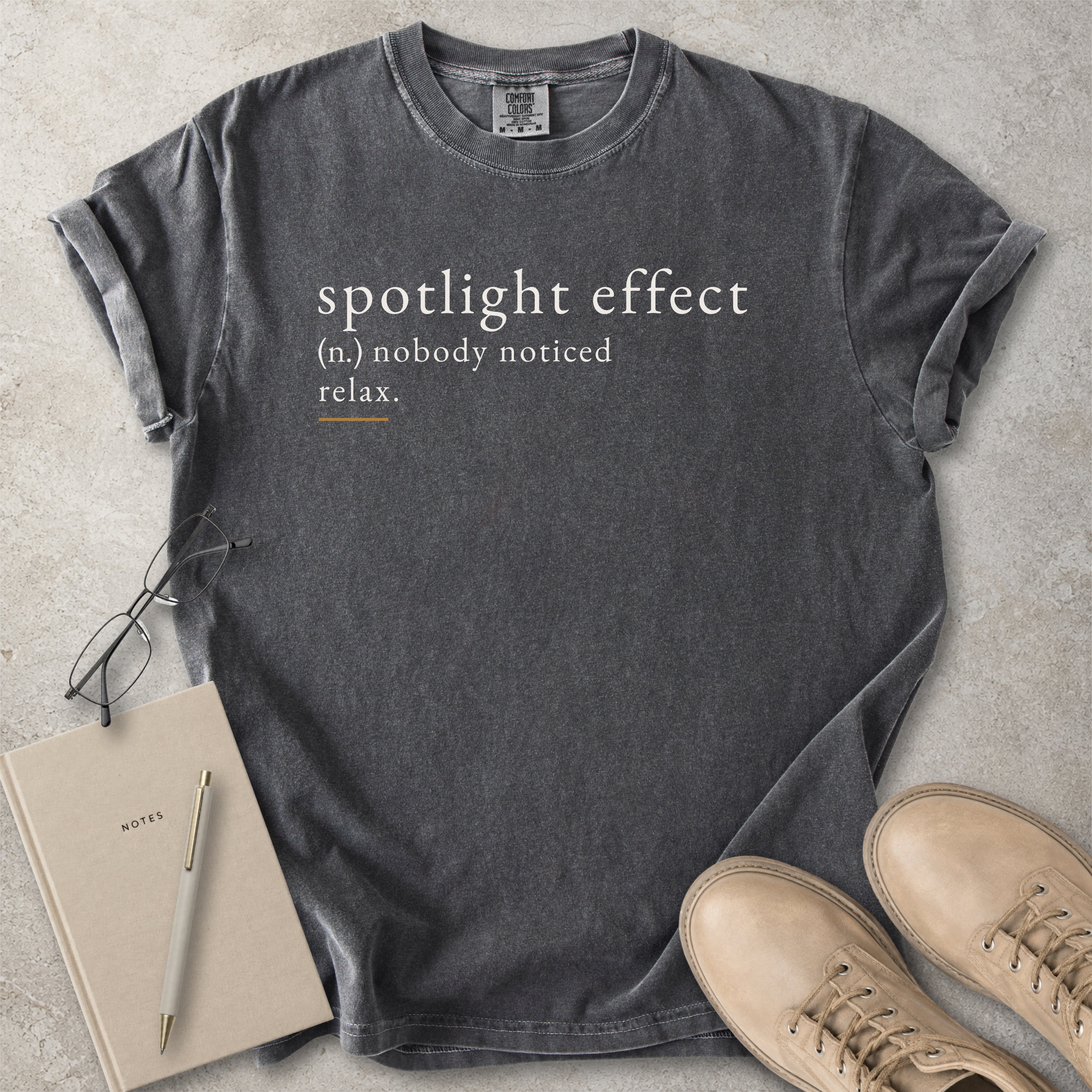 Gray t-shirt with 'spotlight effect' text, notebook, pen, and shoes on a concrete surface
