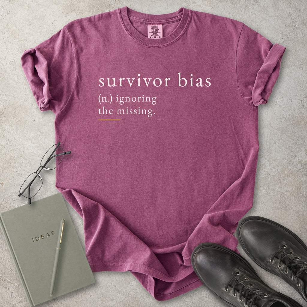 Purple t-shirt with 'survivor bias' text on a concrete surface with glasses, notebook, and shoes.