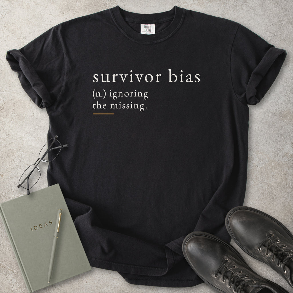 Black t-shirt with 'survivor bias' text on a concrete surface with glasses, notebook, and shoes.