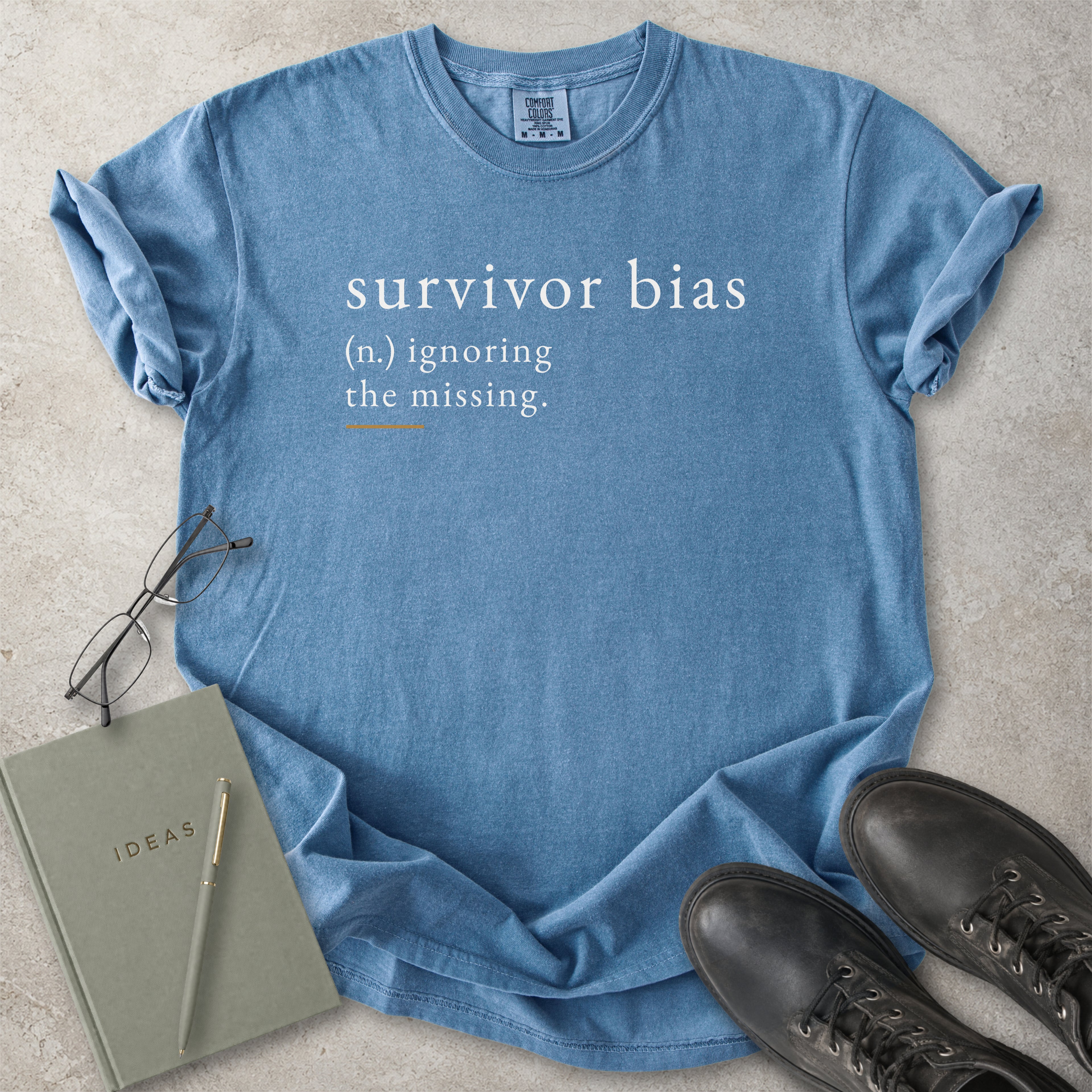 Blue t-shirt with 'survivor bias' text on a concrete surface with glasses, notebook, and shoes.