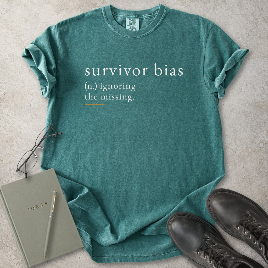 Green t-shirt with 'survivor bias' text on a concrete surface with glasses and notebook.