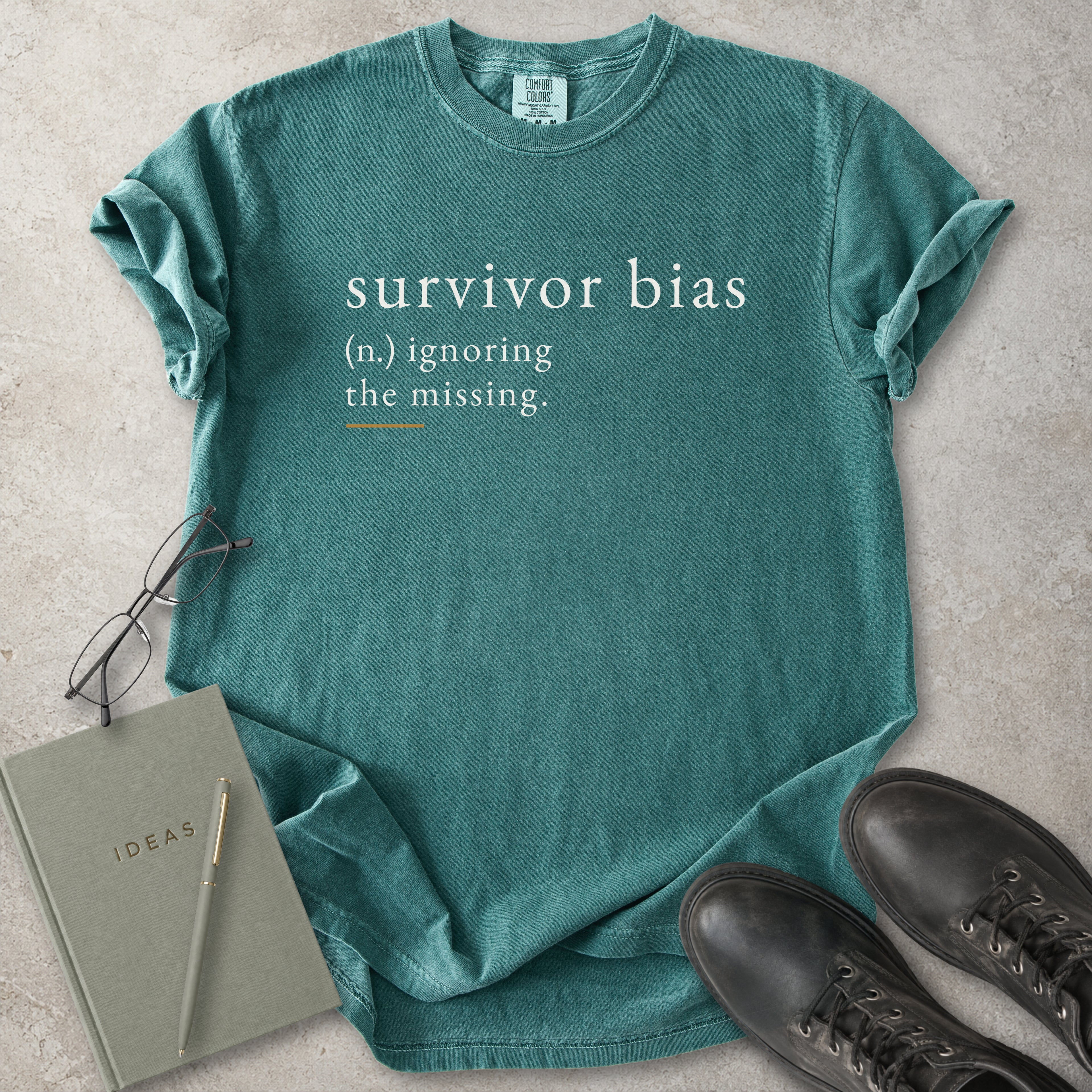 Green t-shirt with 'survivor bias' text on a concrete surface with glasses and notebook.