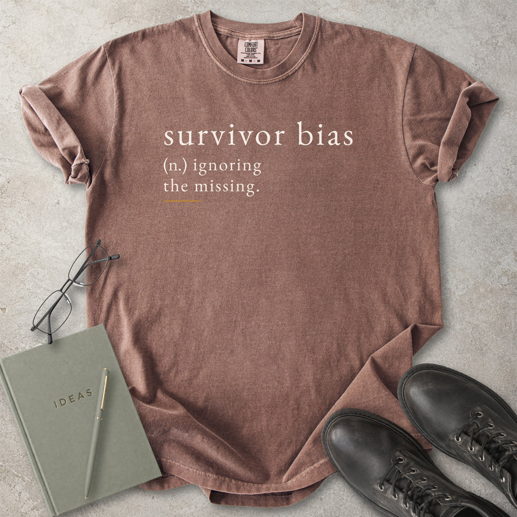 Brown t-shirt with 'survivor bias' text on a concrete surface with glasses, notebook, and shoes.