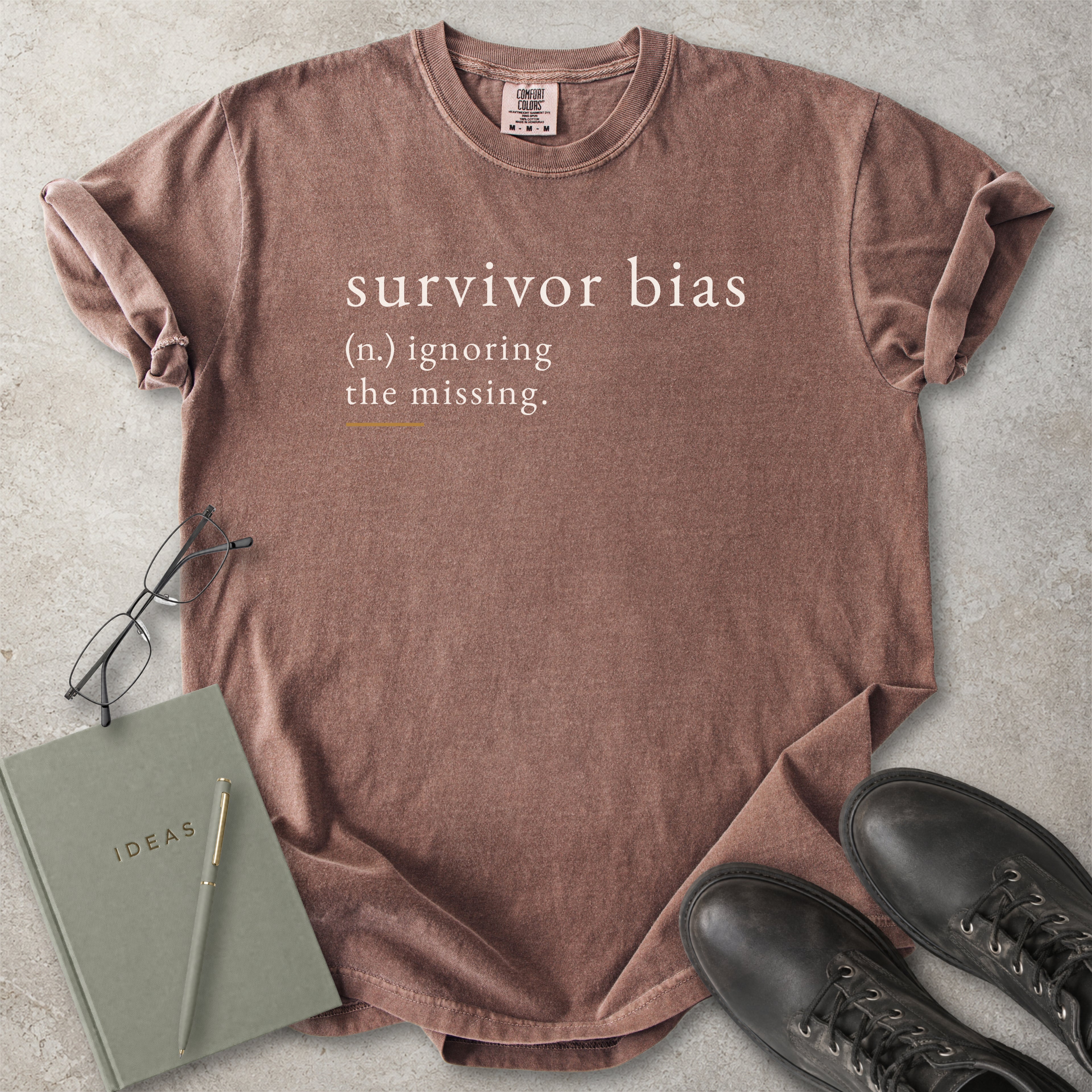 Brown t-shirt with 'survivor bias' text on a concrete surface with glasses, notebook, and shoes.