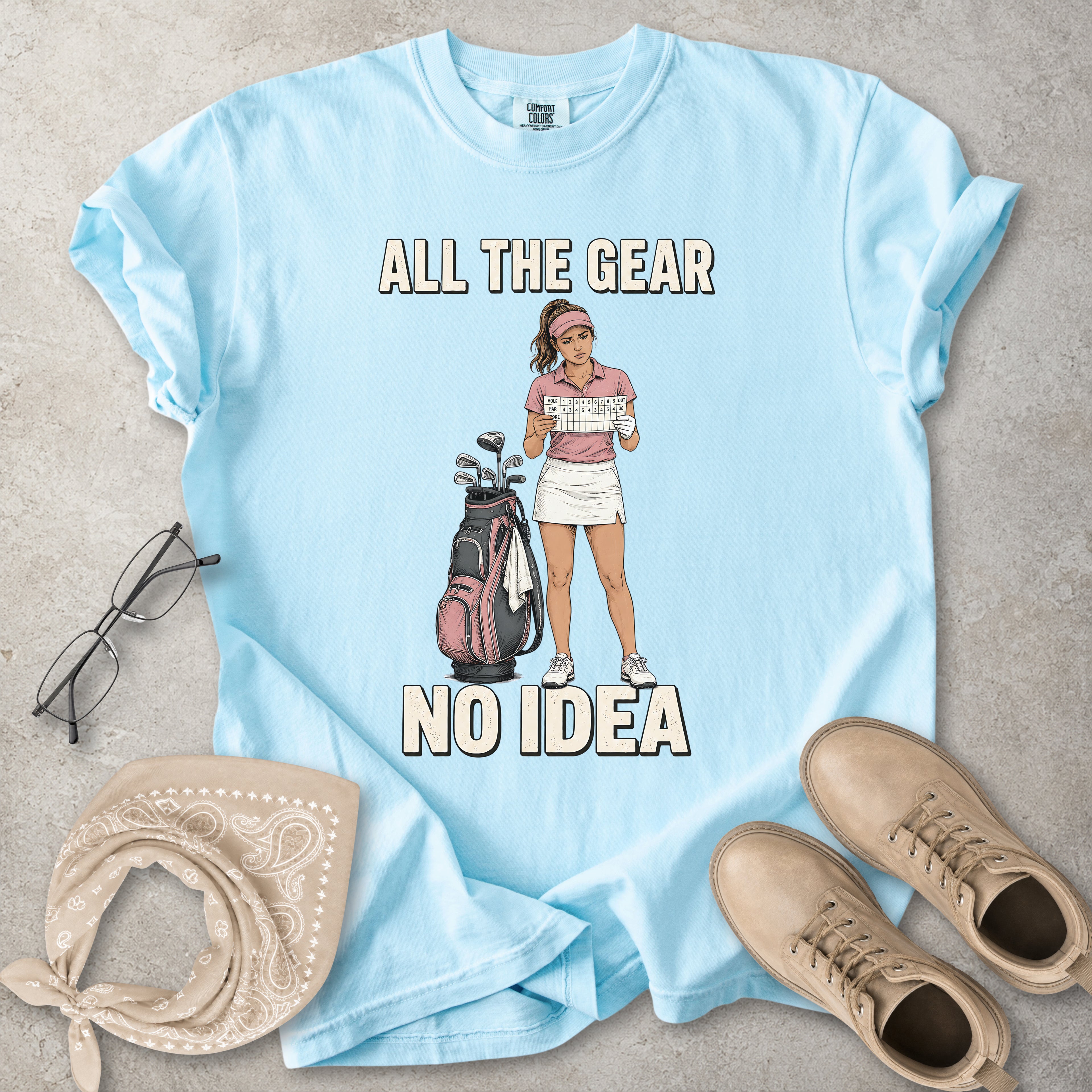 Chambray light blue Comfort Colors 1717 T-shirt with a funny All the Gear No Idea golf graphic showing a woman golfer holding a scorecard beside a golf bag.