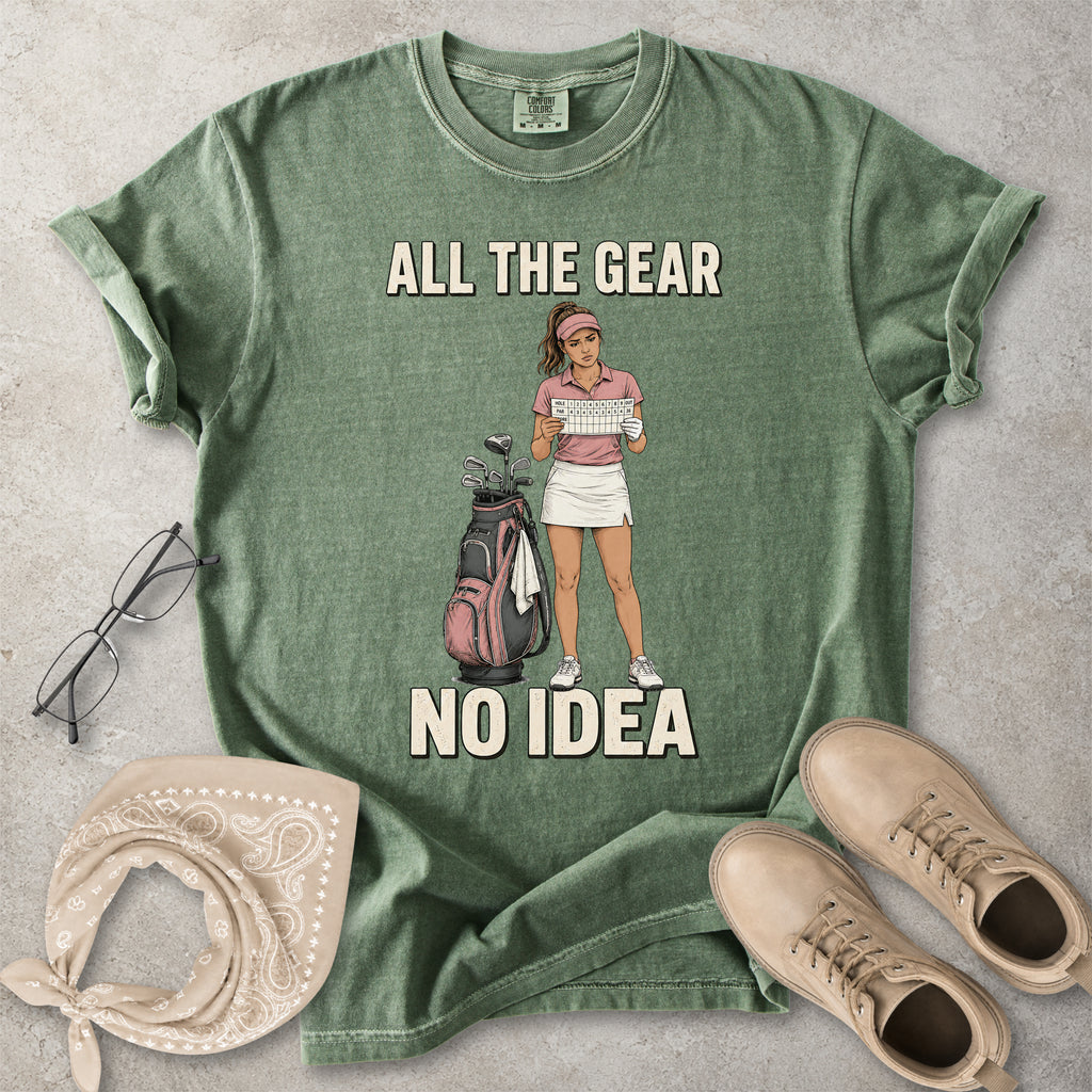 Green t-shirt with graphic of a woman holding a golf club and text 'All the gear, no idea' on a concrete surface with accessories.