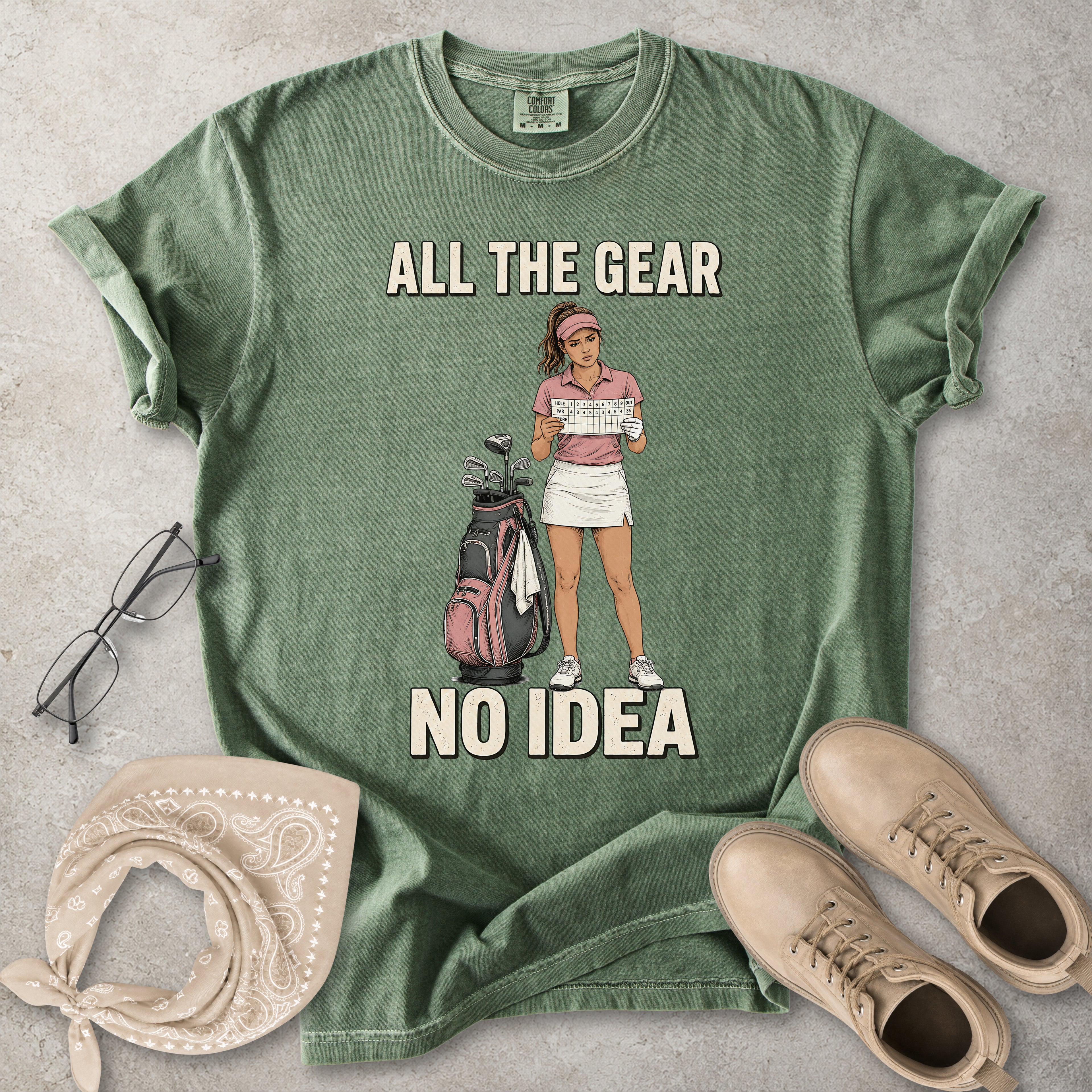 Green t-shirt with graphic of a woman holding a golf club and text 'All the gear, no idea' on a concrete surface with accessories.