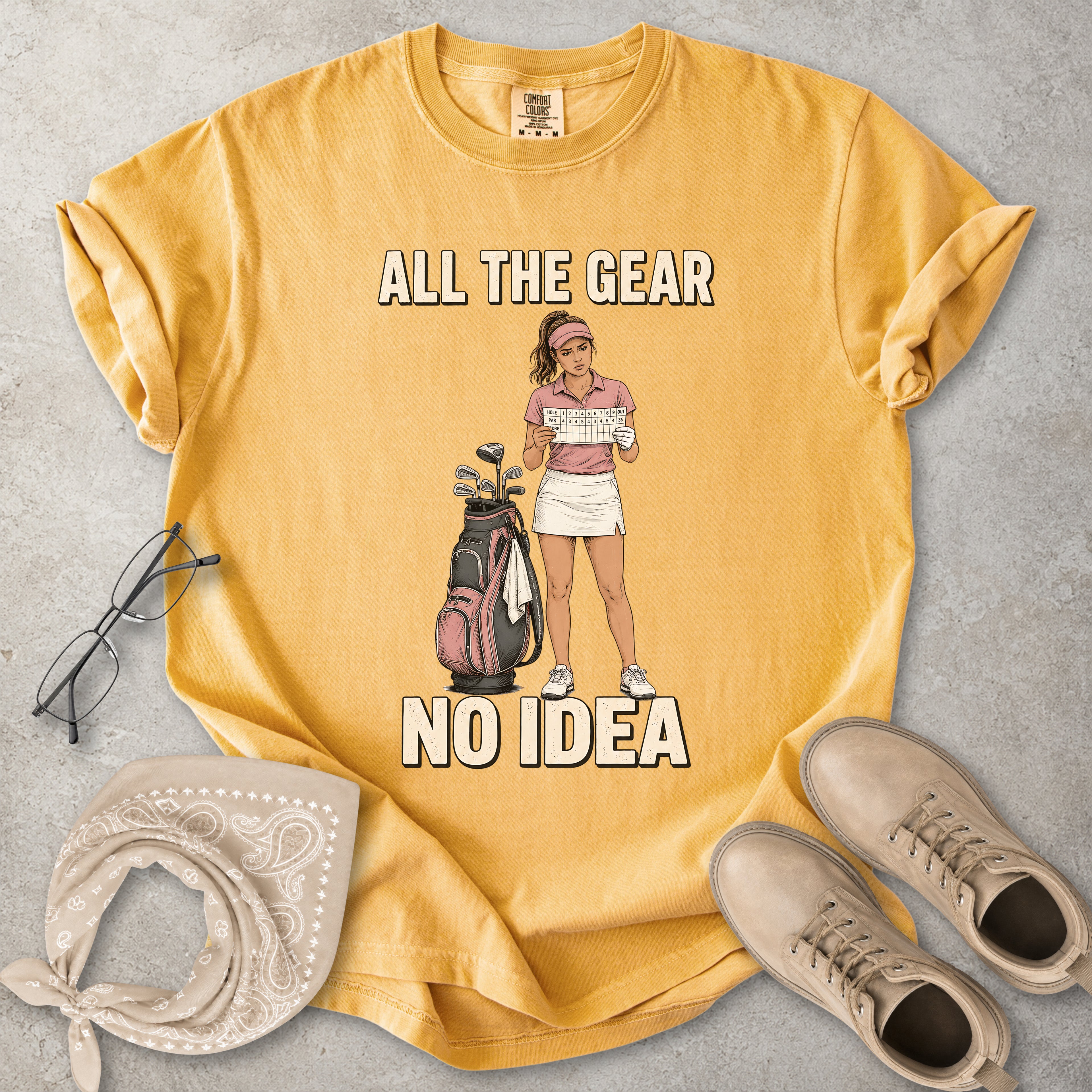 Yellow t-shirt with graphic design of a woman holding a golf club and text 'All the gear, no idea' on a gray surface with accessories.