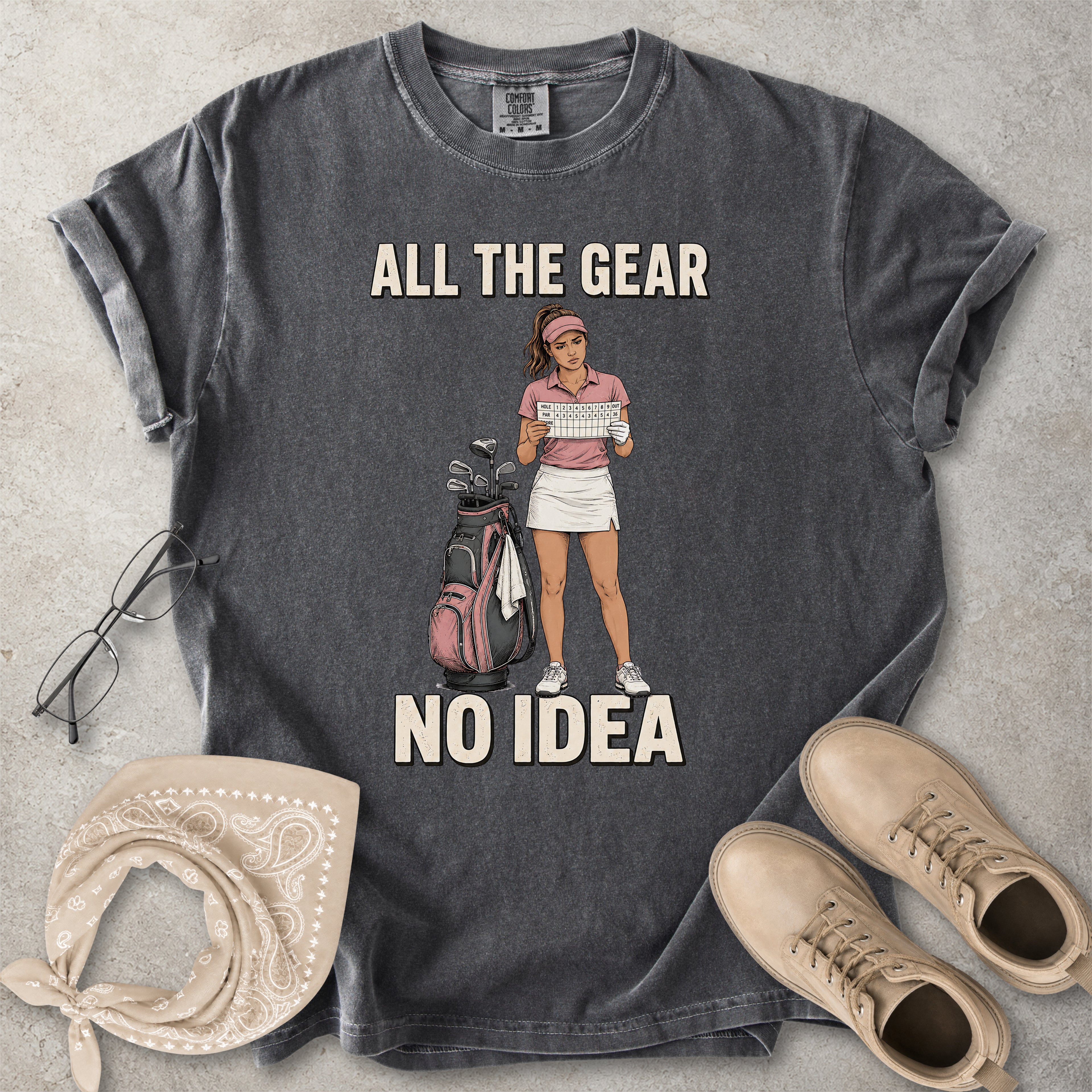 Gray t-shirt with graphic of a woman holding golf clubs and text 'All the gear, no idea' on a concrete surface with accessories.